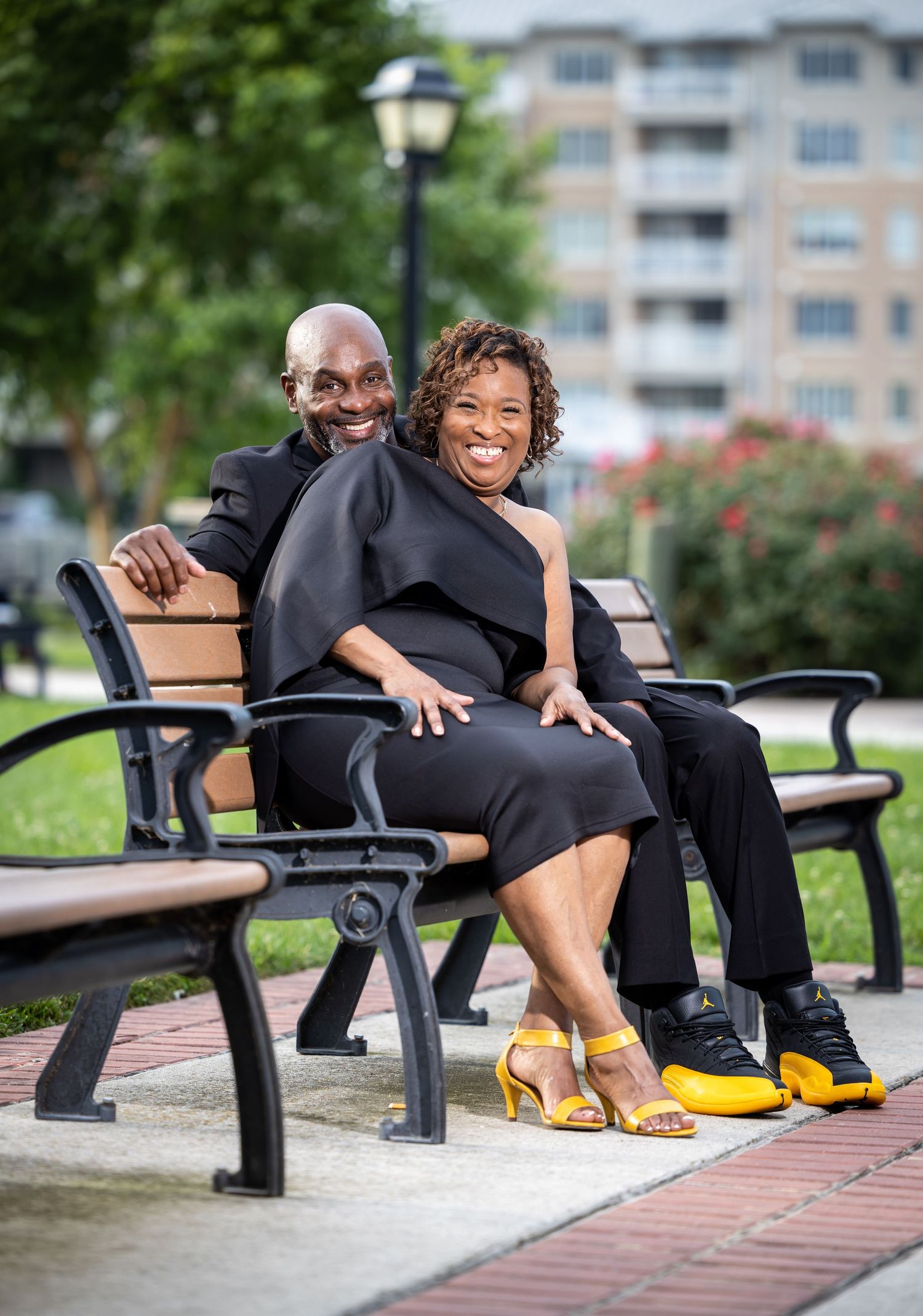 A couple shares a cheerful moment while sitting on a park bench, wearing coordinated black formal attire.
