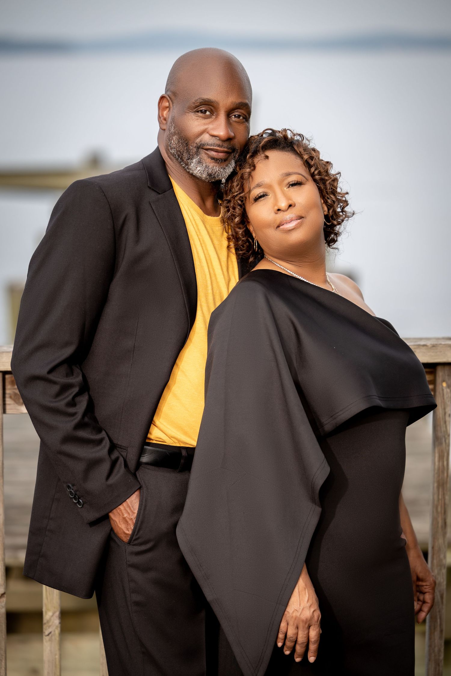 A couple in formal wear poses together, with the person in yellow shirt and black blazer standing behind.