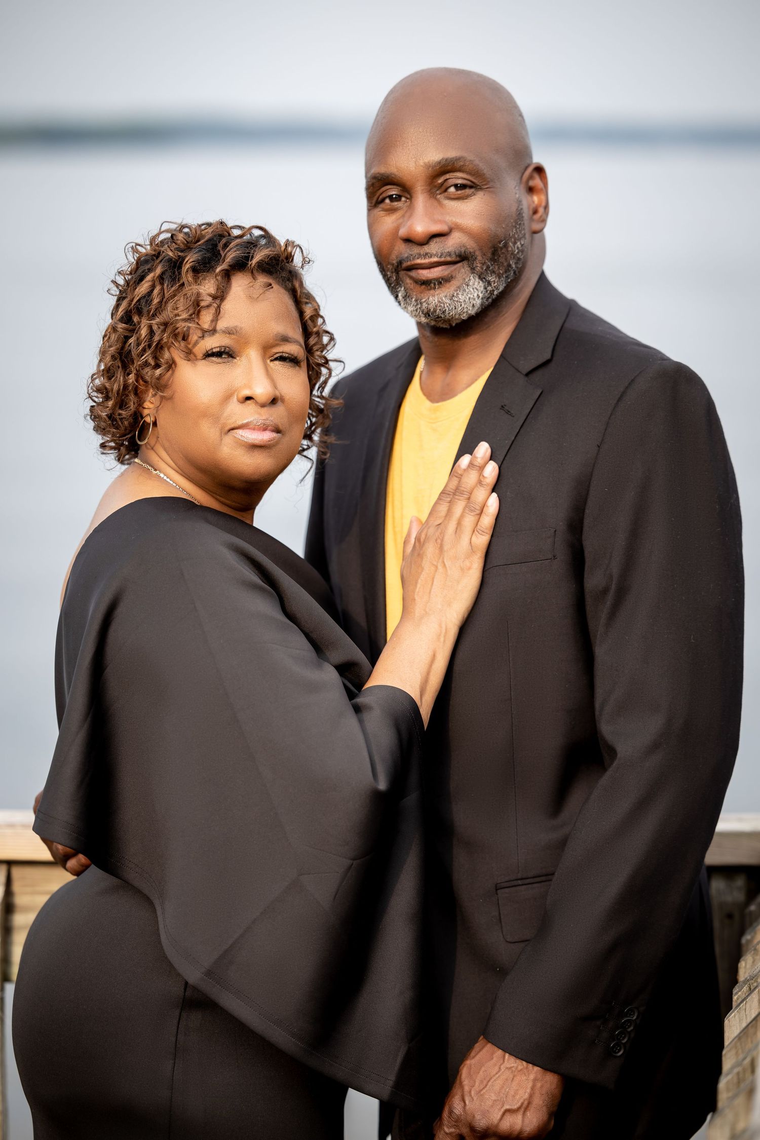 A couple in coordinated black and yellow formal wear poses together against a waterfront backdrop.