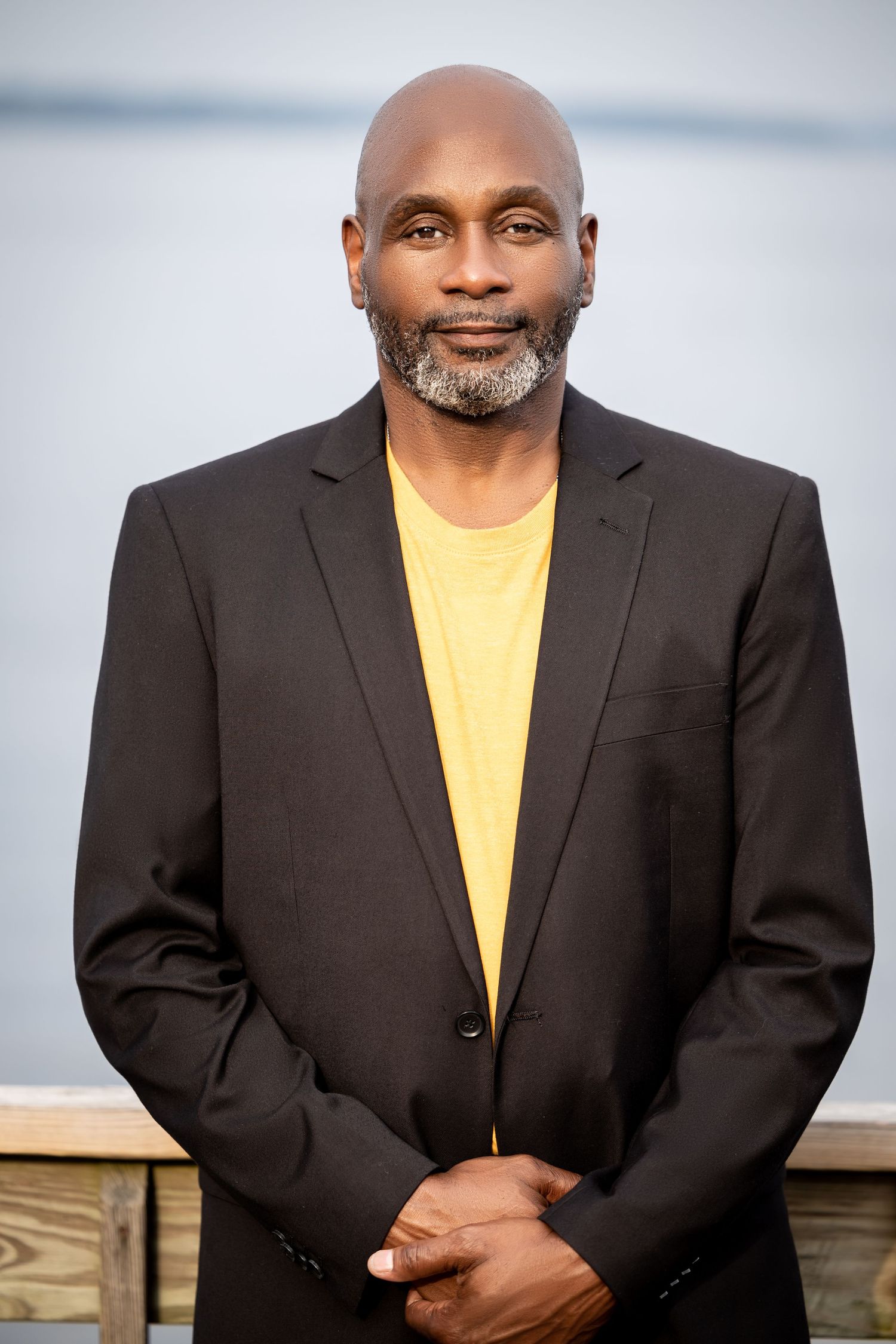 Portrait of a person in a black blazer and yellow shirt with a neatly trimmed beard against a waterfront backdrop.