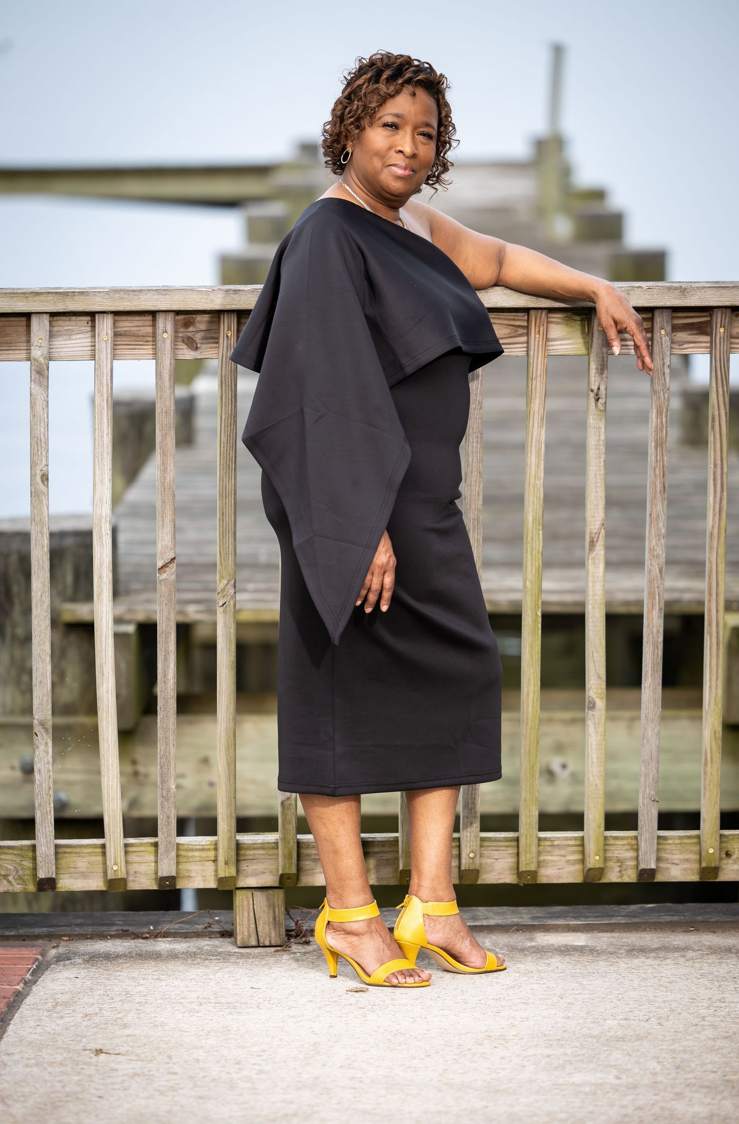 Person in a black dress and yellow heels poses elegantly against a deck railing.