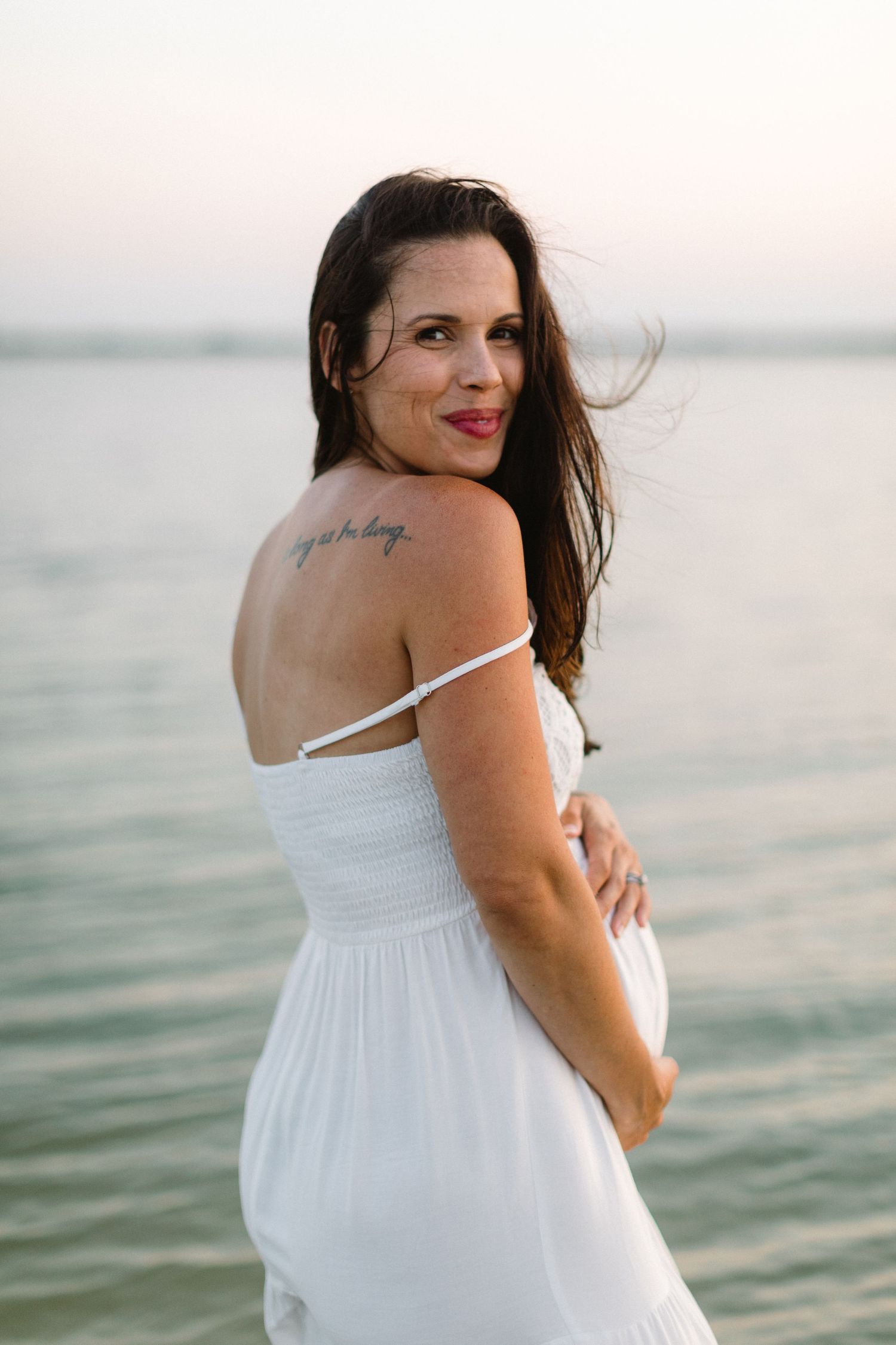 A person in a white dress poses by the water at sunset, showing a tattoo on their shoulder.