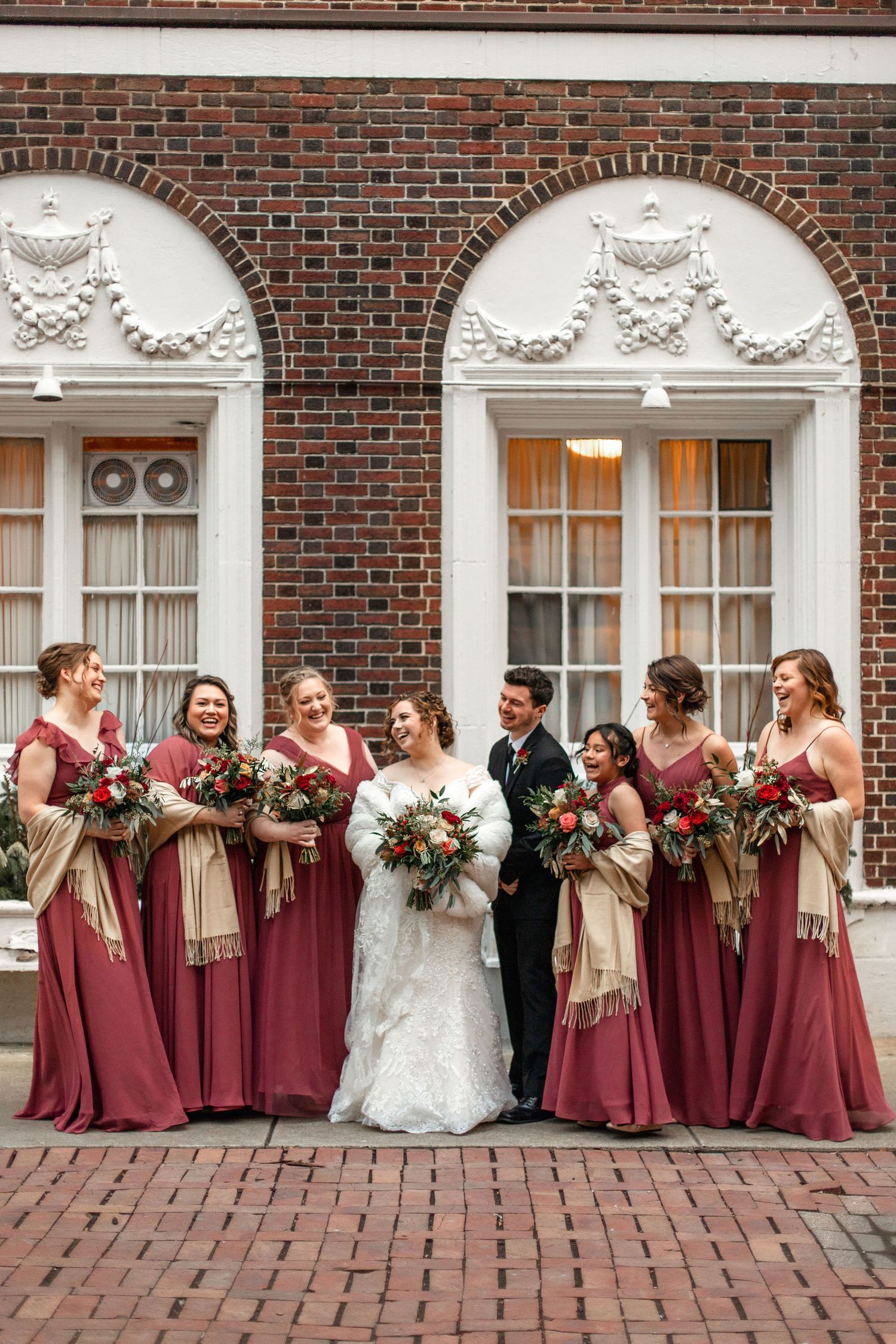 Wedding party in burgundy dresses poses in front of historic brick building with ornate white archways.