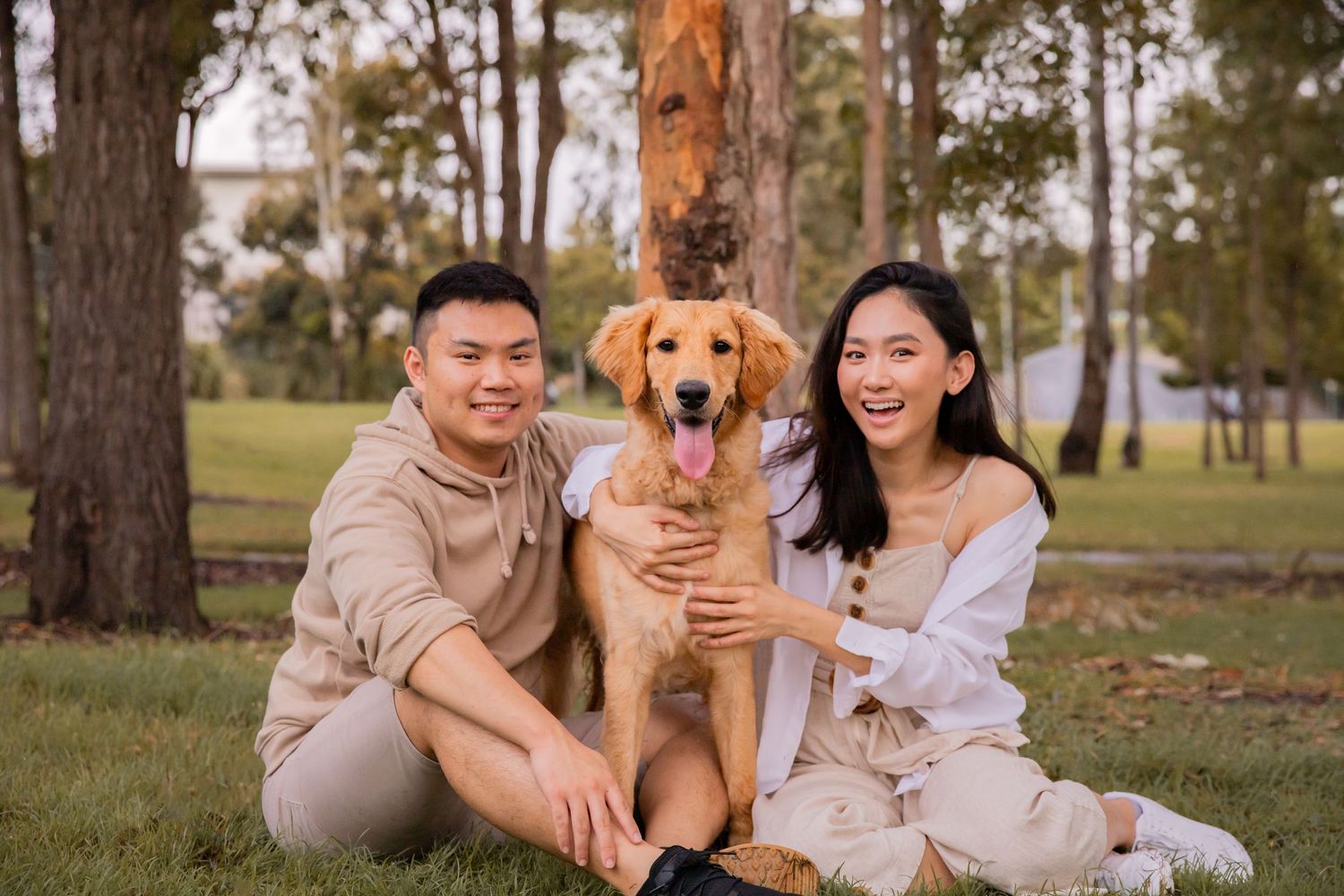 Two people sit on the ground with a Goldendoodle between them, all smiling and looking at the camera