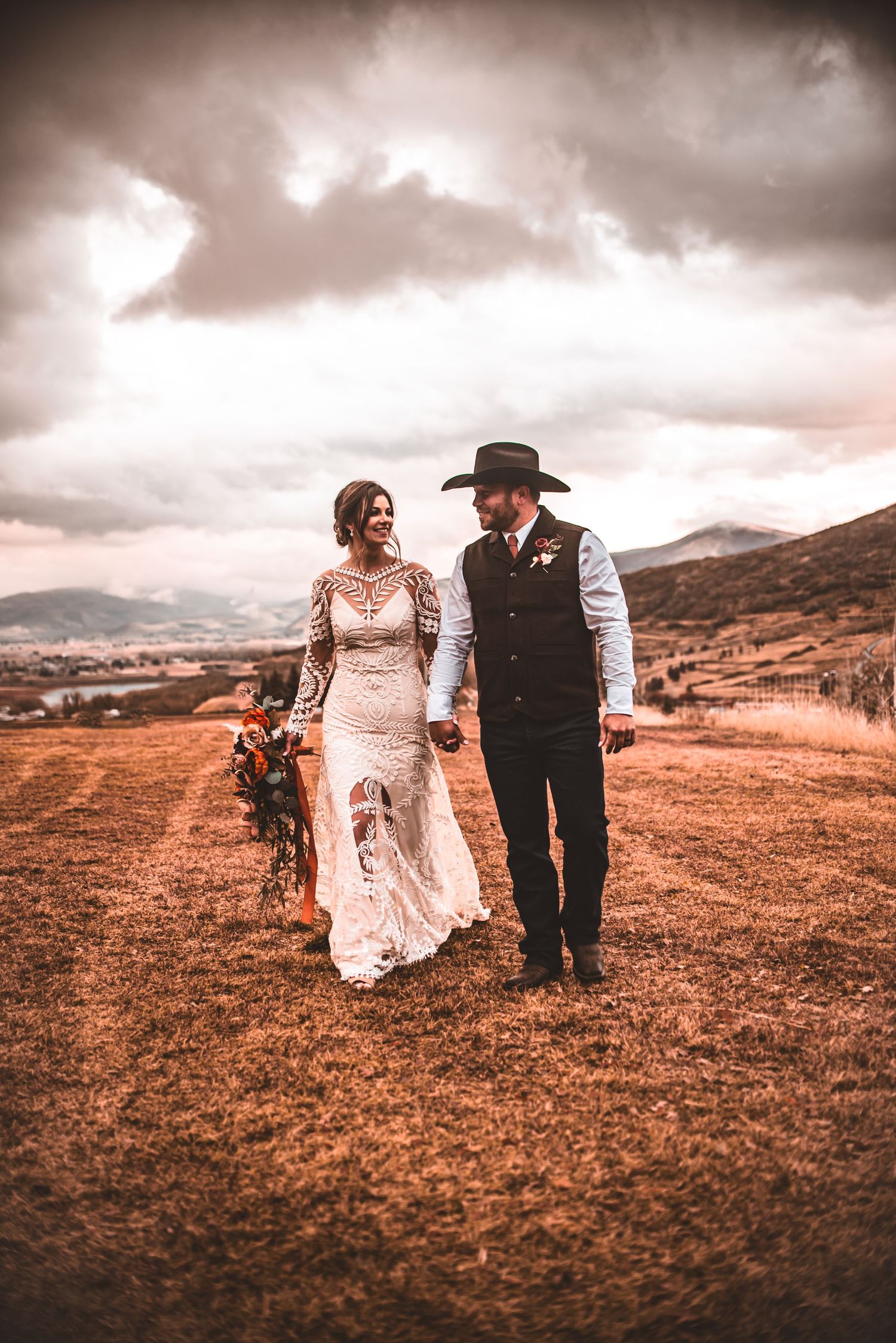 A wedding couple strolls across a hilltop under dramatic sunset clouds in western attire.
