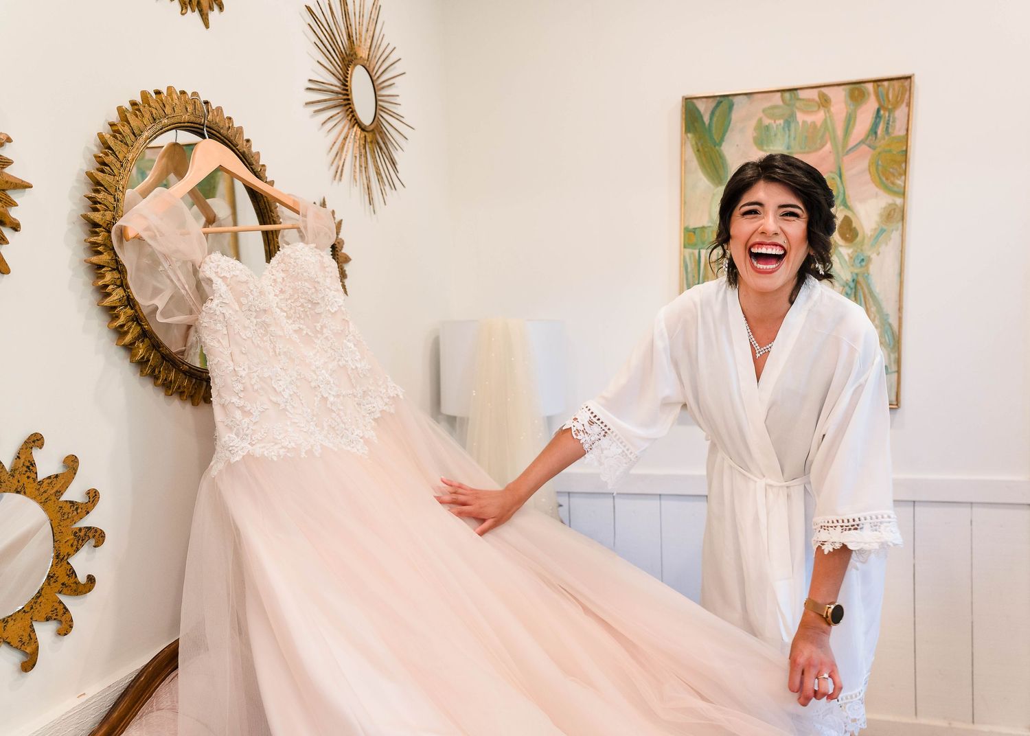 A wedding dress displayed on wall with decorative mirrors while someone celebrates beside it.