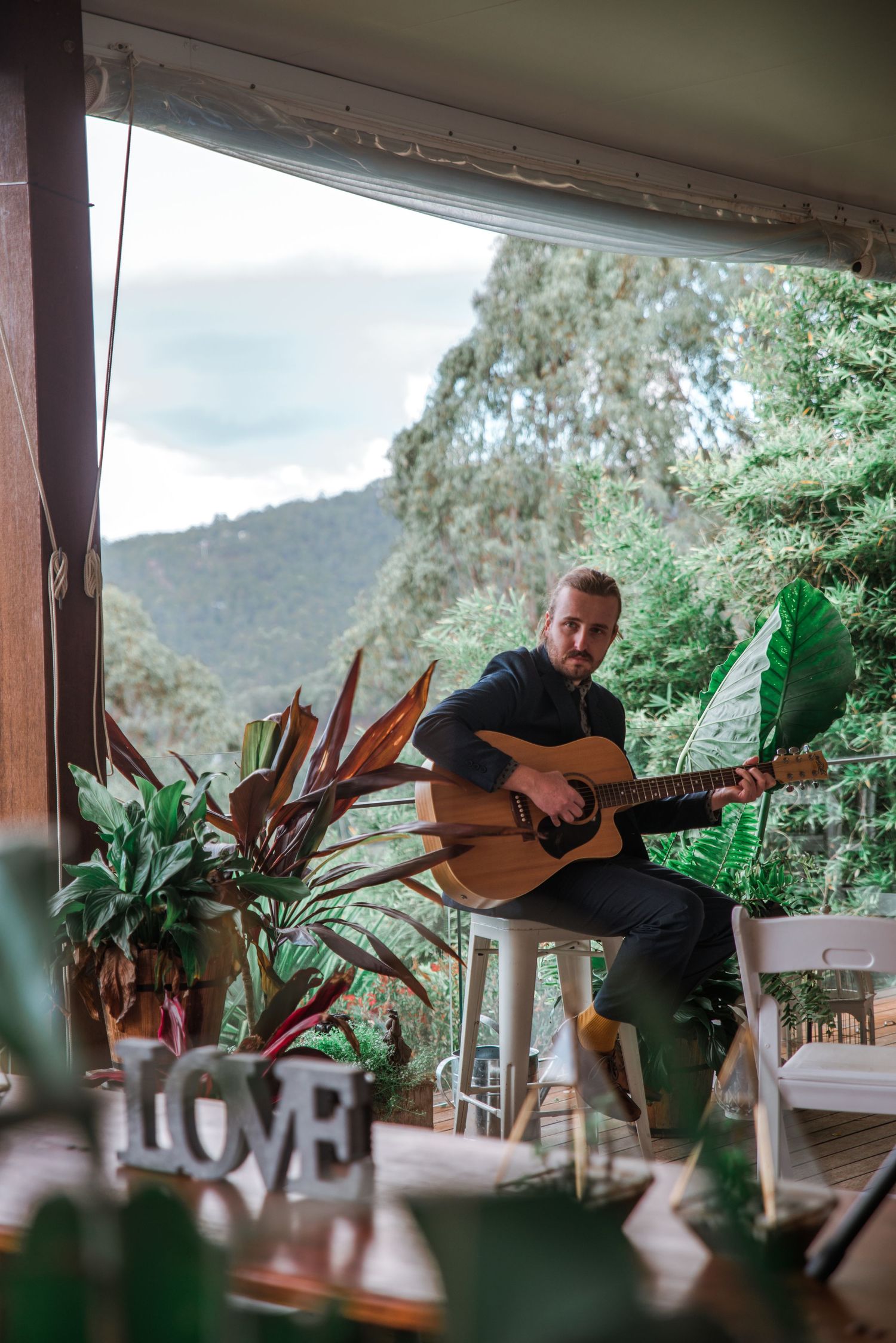 Musician performing with acoustic guitar on covered porch with mountain backdrop.