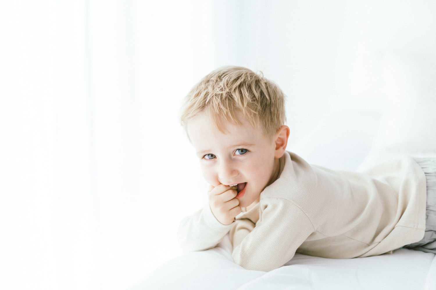 Child in beige sweater laying on white surface with playful expression.