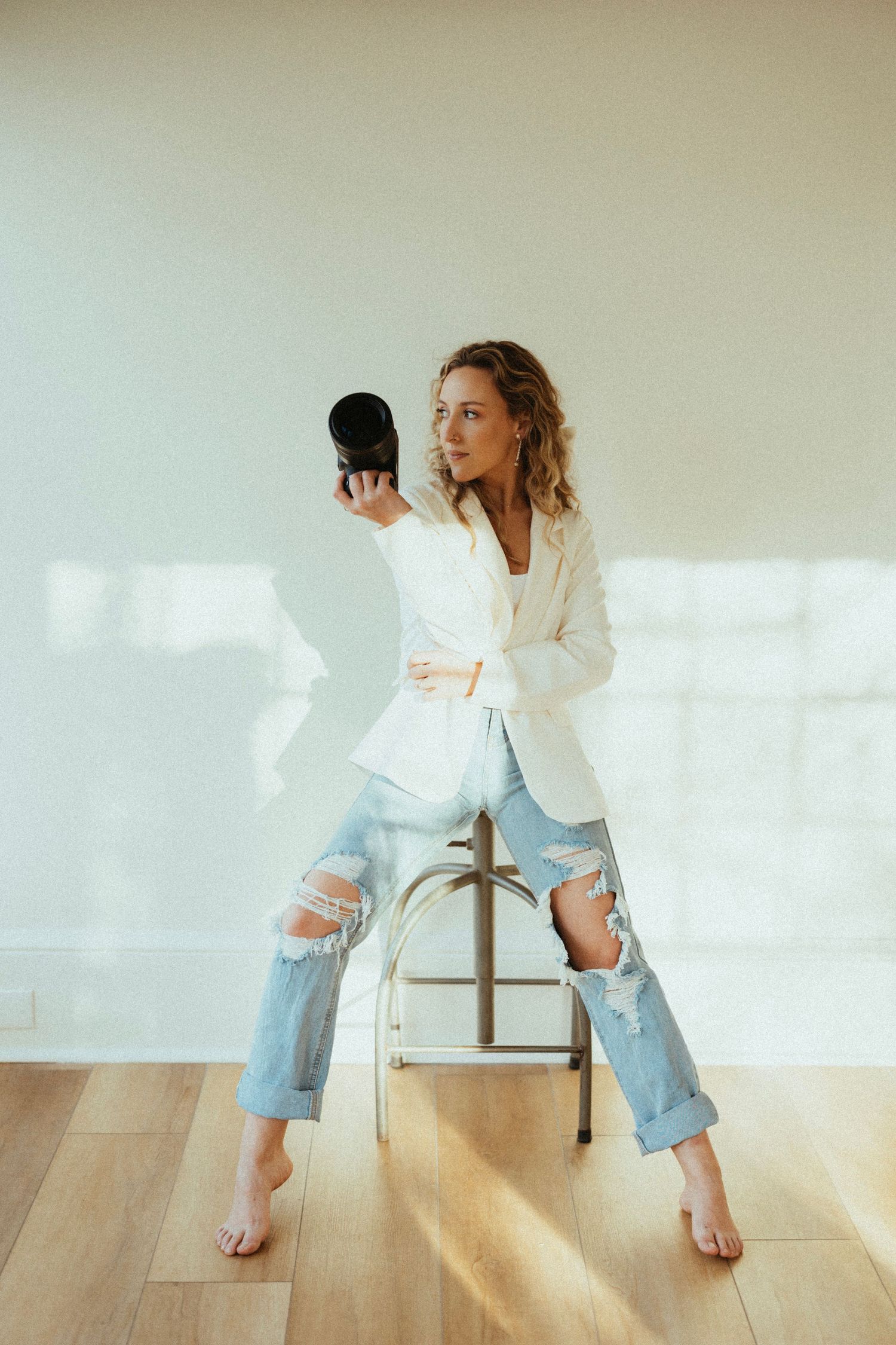 Photographer in white blazer and distressed jeans holding camera in bright studio setting.