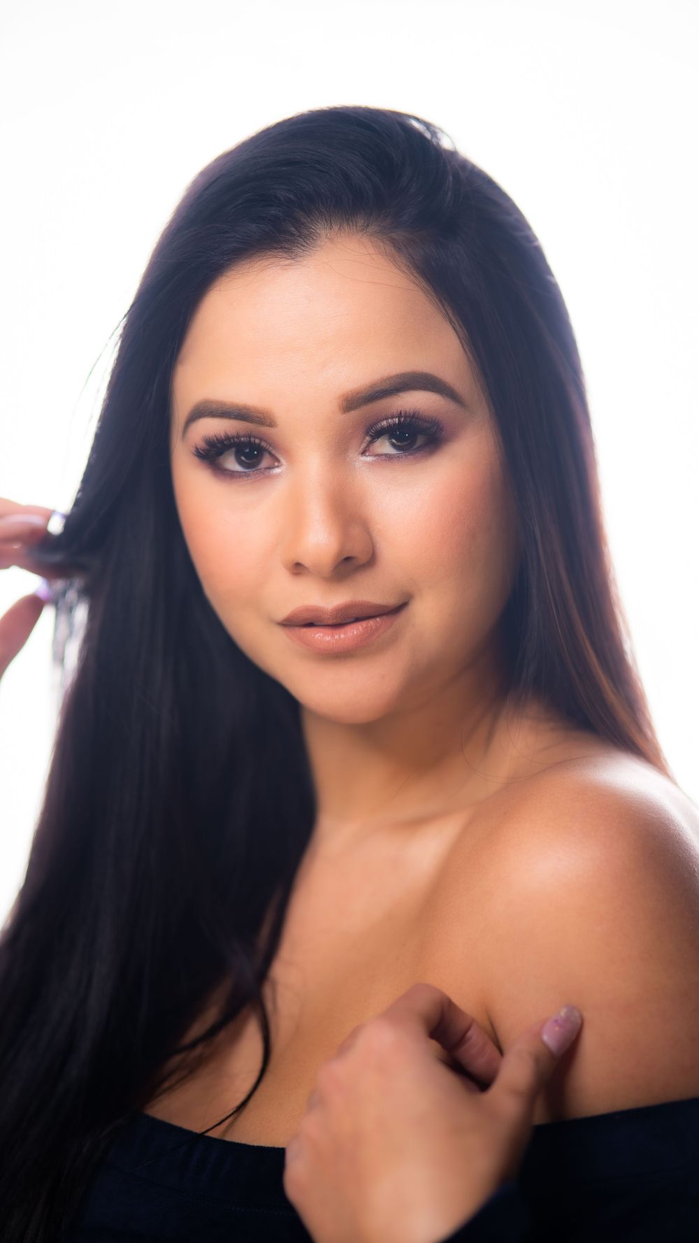 Young woman with long black hair poses confidently in an off-shoulder top against a white background.