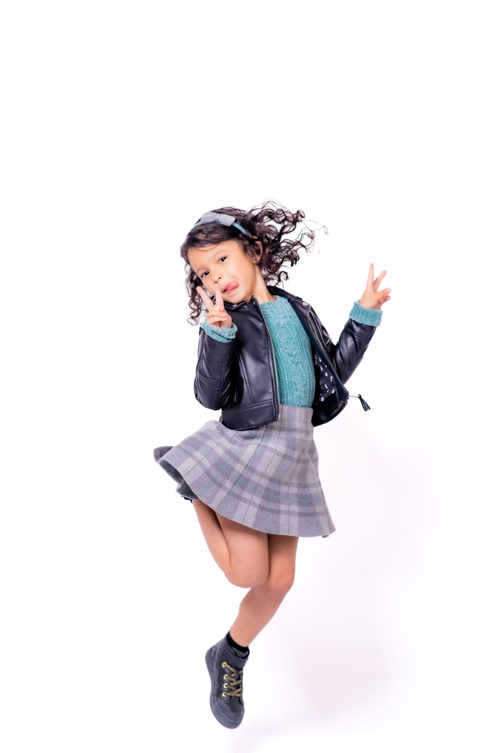 Energetic little girl jumps playfully wearing a leather jacket, plaid skirt, and boots.