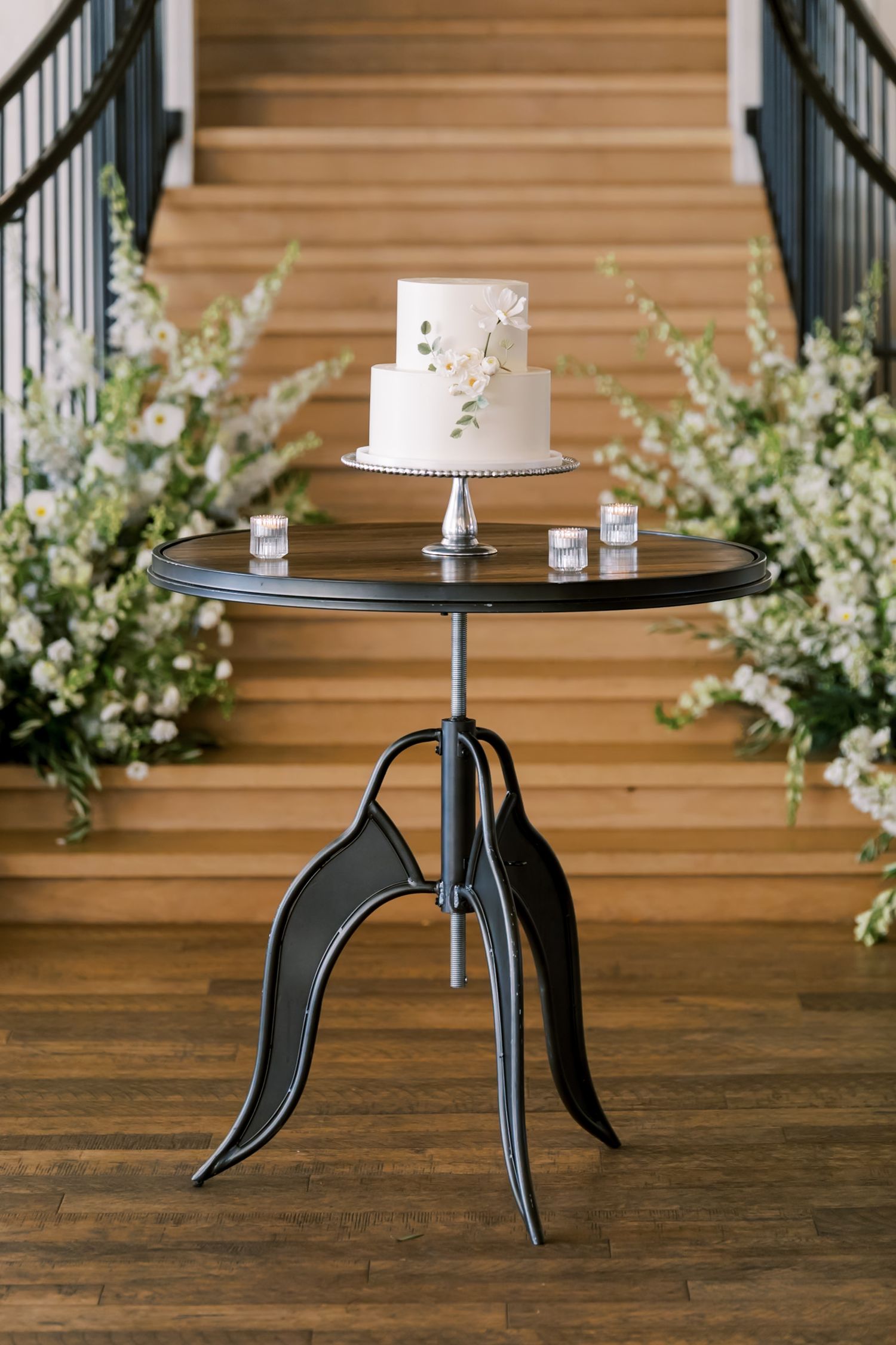 Simple white wedding cake displayed on modern metal stand with floral decorations.