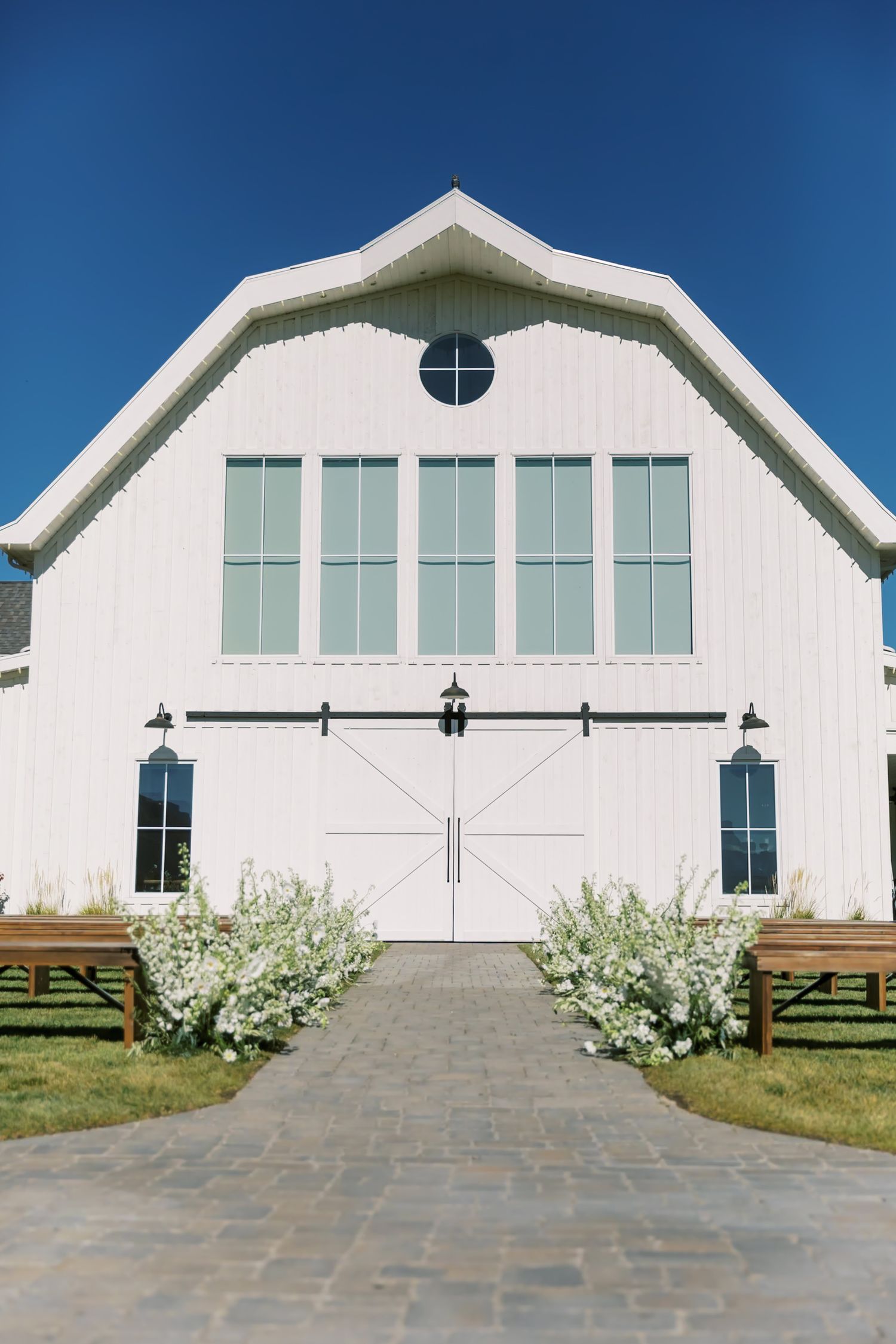 White barn wedding venue with elegant doors, round window and flower-lined entrance path.