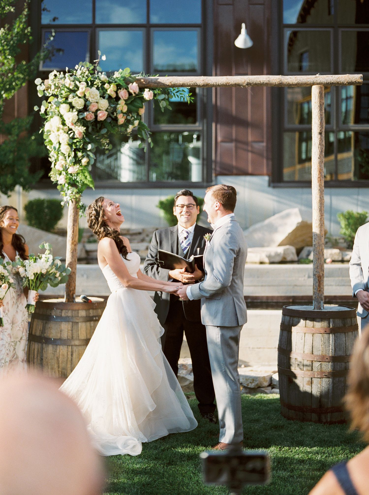 A rustic outdoor wedding ceremony under a wooden arch decorated with white floral arrangements and wine barrels.