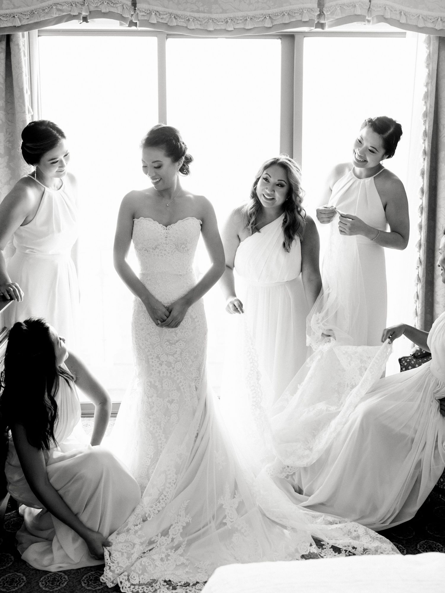 Ladies in white dresses share a joyful moment while getting ready in a sun-lit bridal suite.