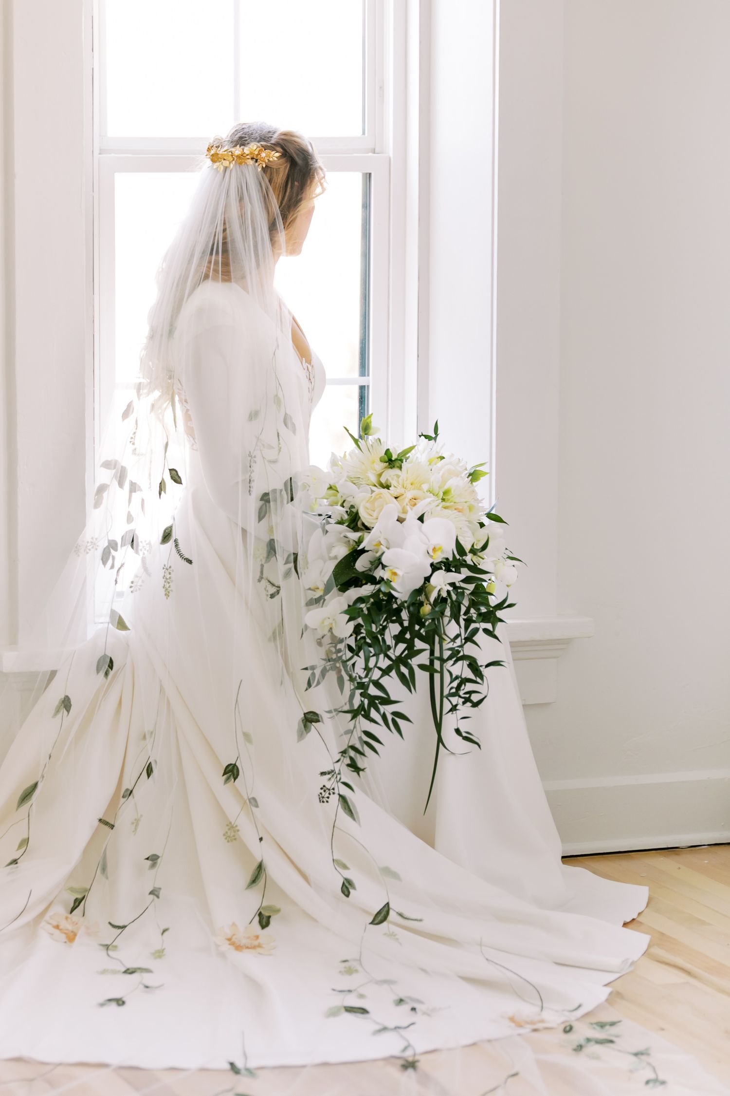 Elegant bride in a floral-detailed wedding gown stands by window with white lily bouquet.