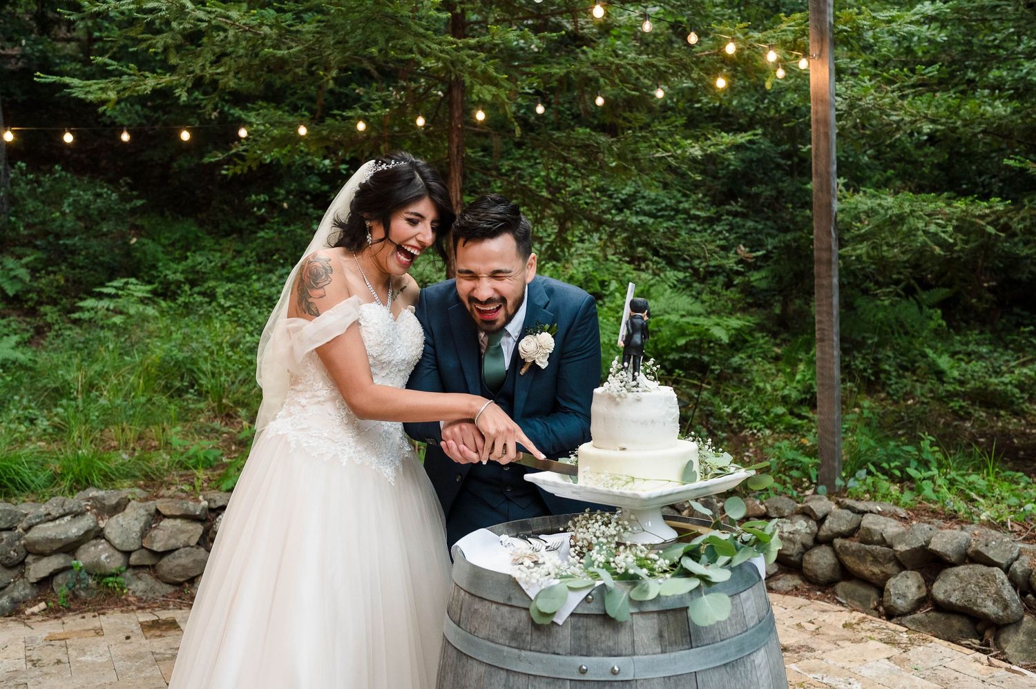 A couple cuts their wedding cake together on a rustic barrel with string lights in the background.