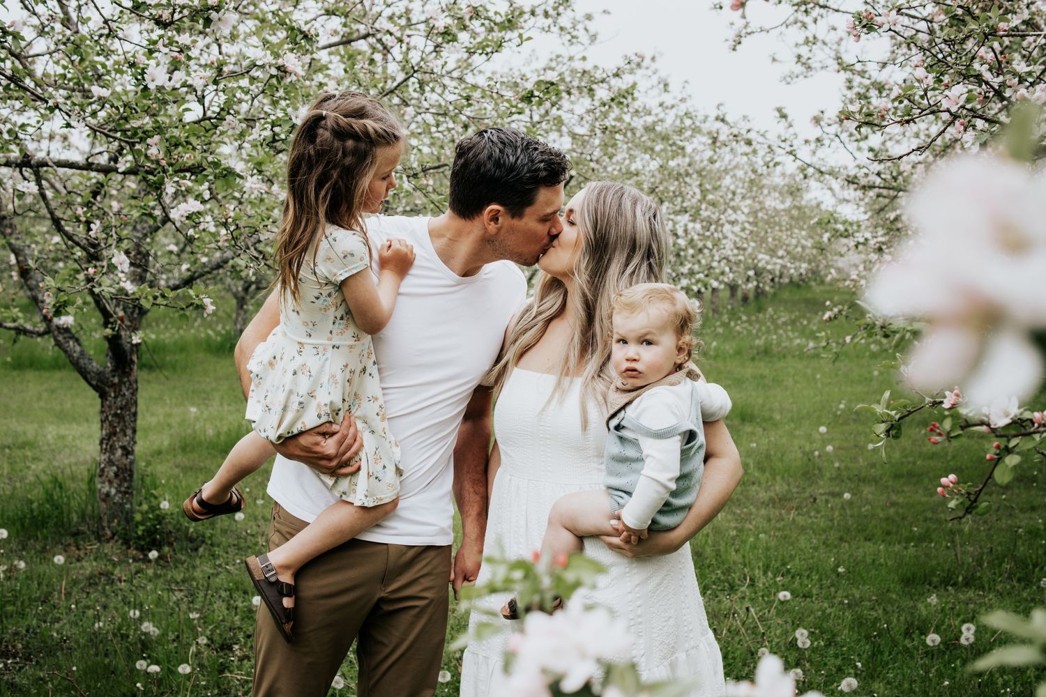A tender family moment captured in a blooming spring orchard with white flowers and green grass.