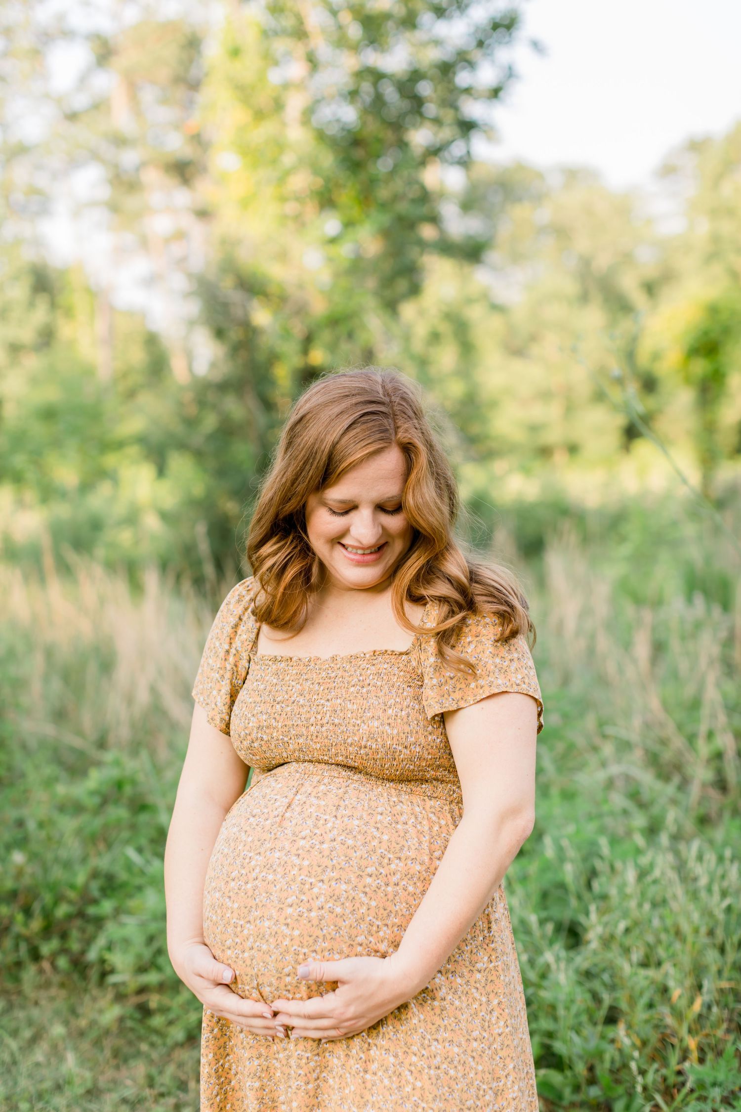 Woman in yellow floral dress looks down at baby bump while standing in nature.
