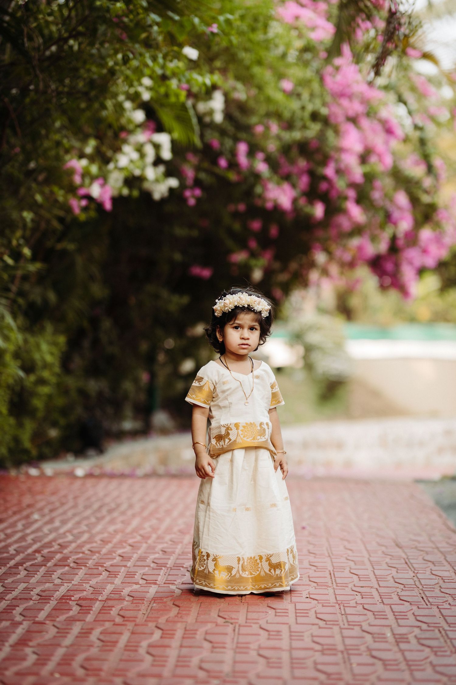 Small child in white and gold traditional dress standing on brick pathway with pink flowers above.