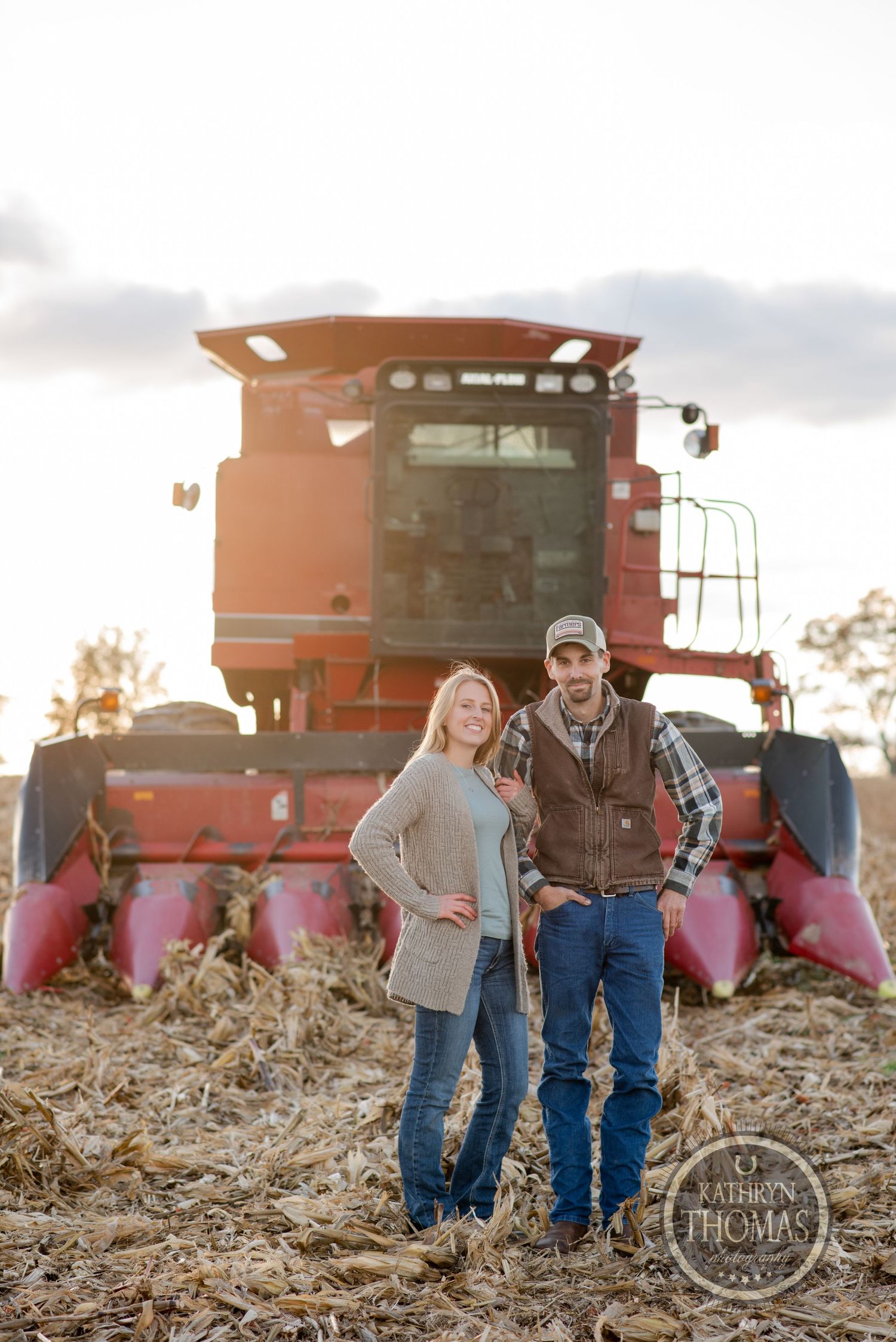 Couple posing in front of large red combine harvester at sunset.