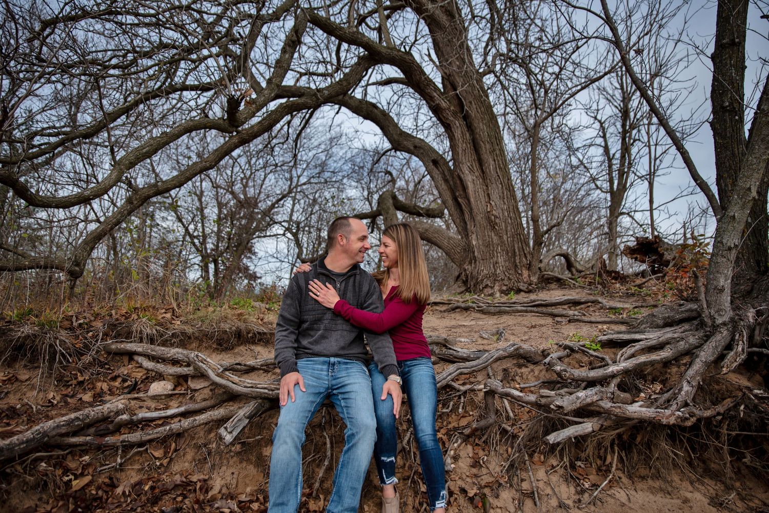 A couple sits together on exposed tree roots in a wooded area during an outdoor photo session.