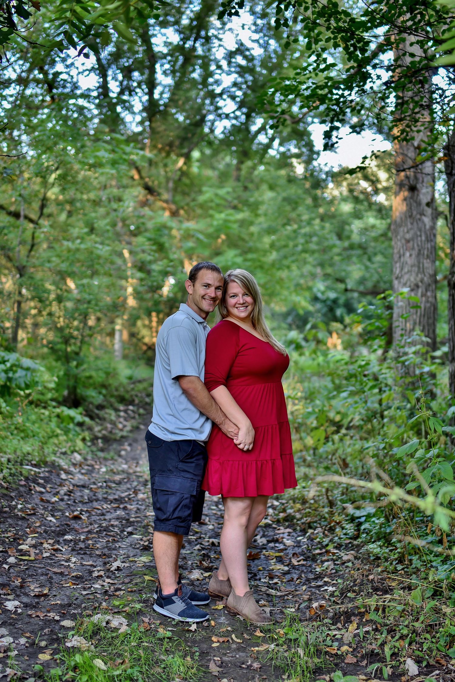 A couple poses on a wooded path, with one wearing red dress and other in casual attire.