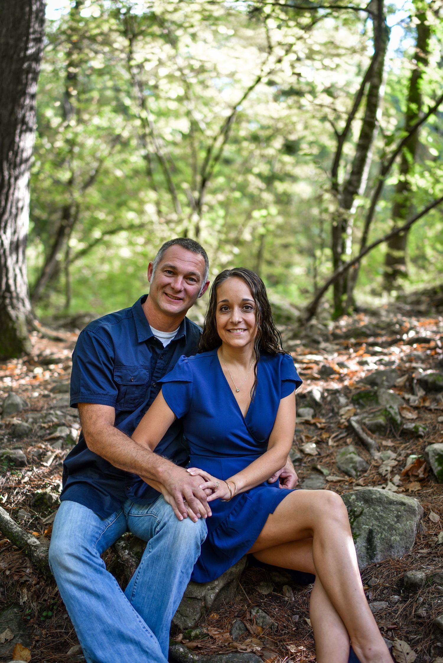 A couple in matching blue outfits sitting together on rocks in a wooded forest setting.