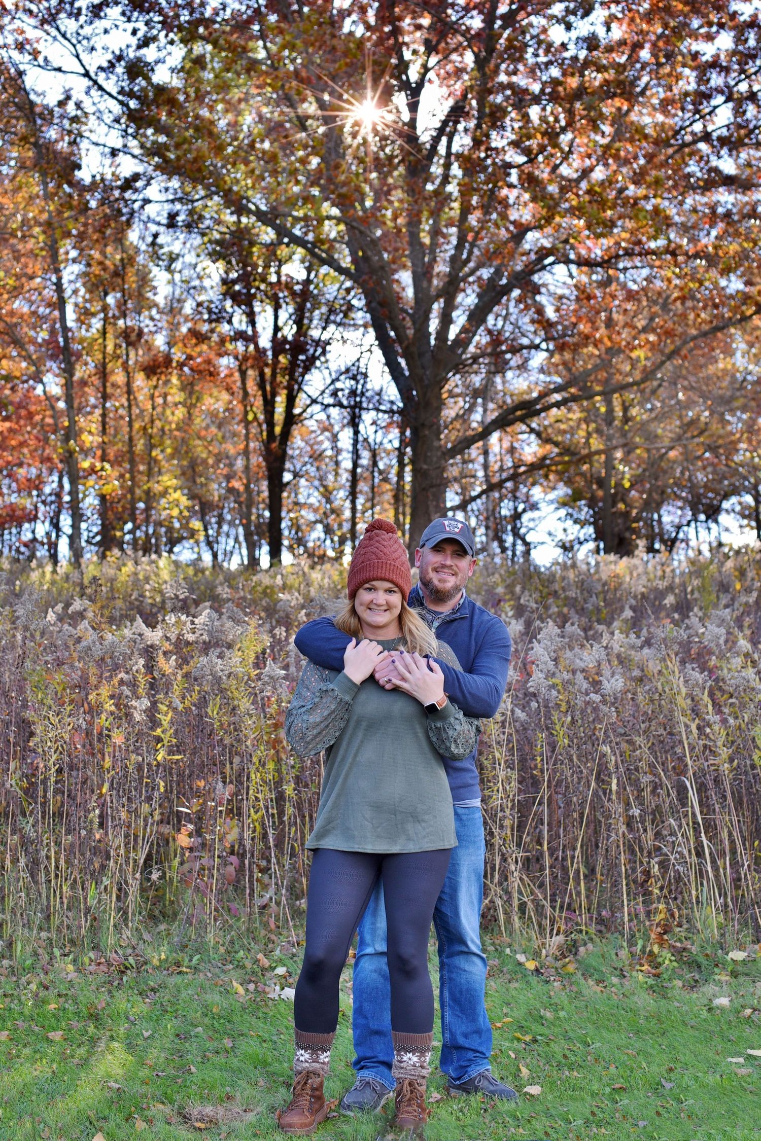 A cozy fall portrait of a couple embracing in front of autumn foliage with sunburst through trees.