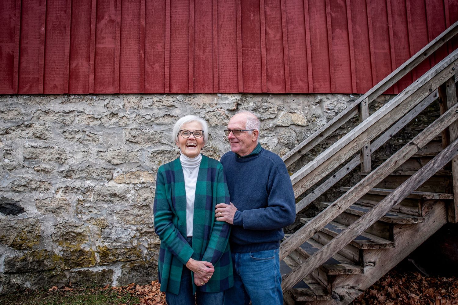 Couple standing by wooden stairs against red barn wall on cloudy day.