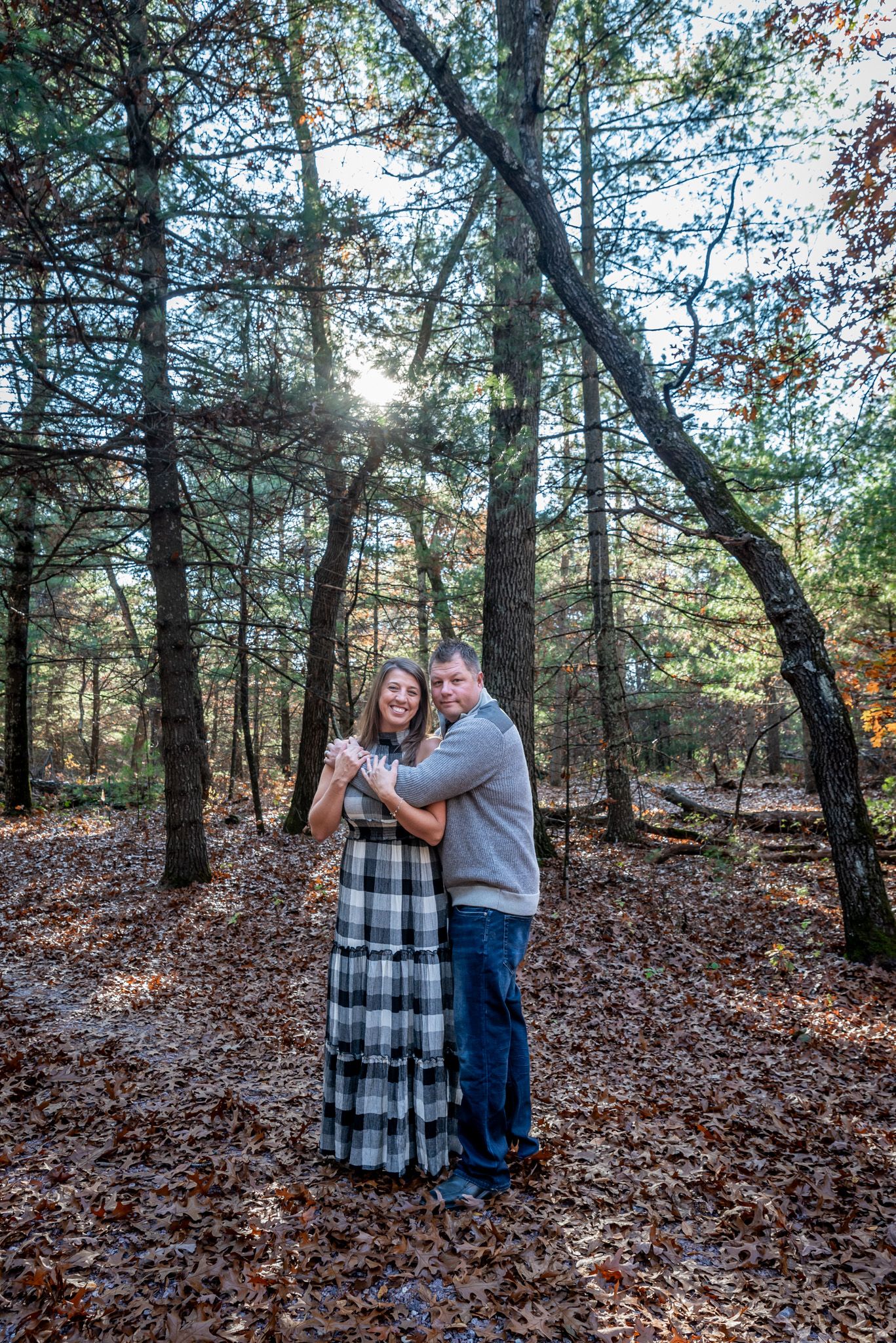 Couple embracing in autumn forest with sunlight filtering through pine trees.