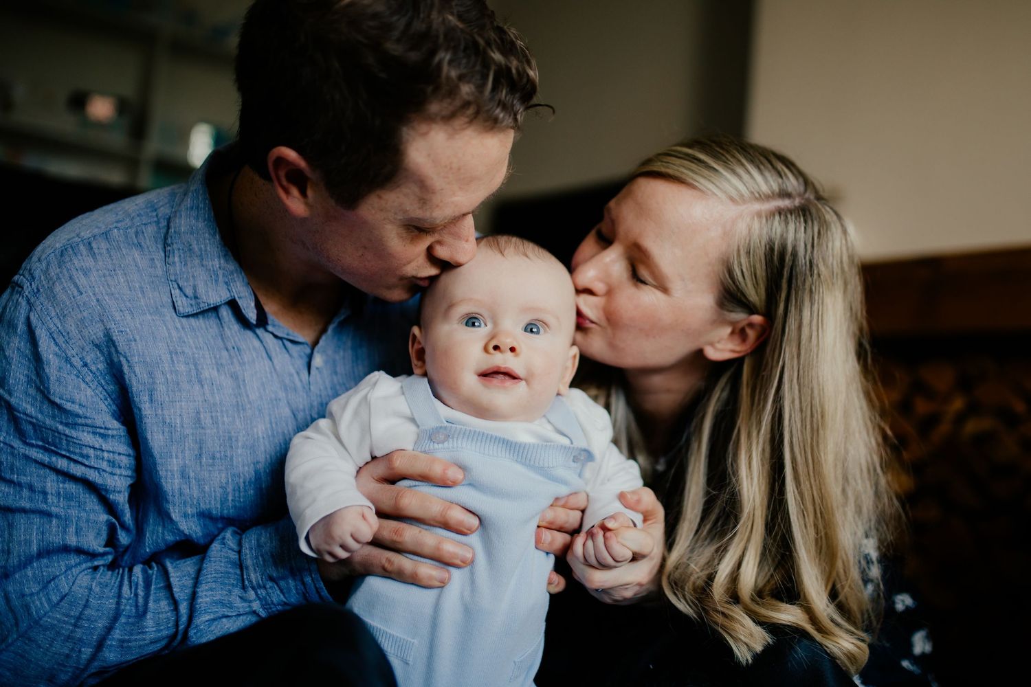 Mother and father sit on the floor and kiss their baby at home in their living room