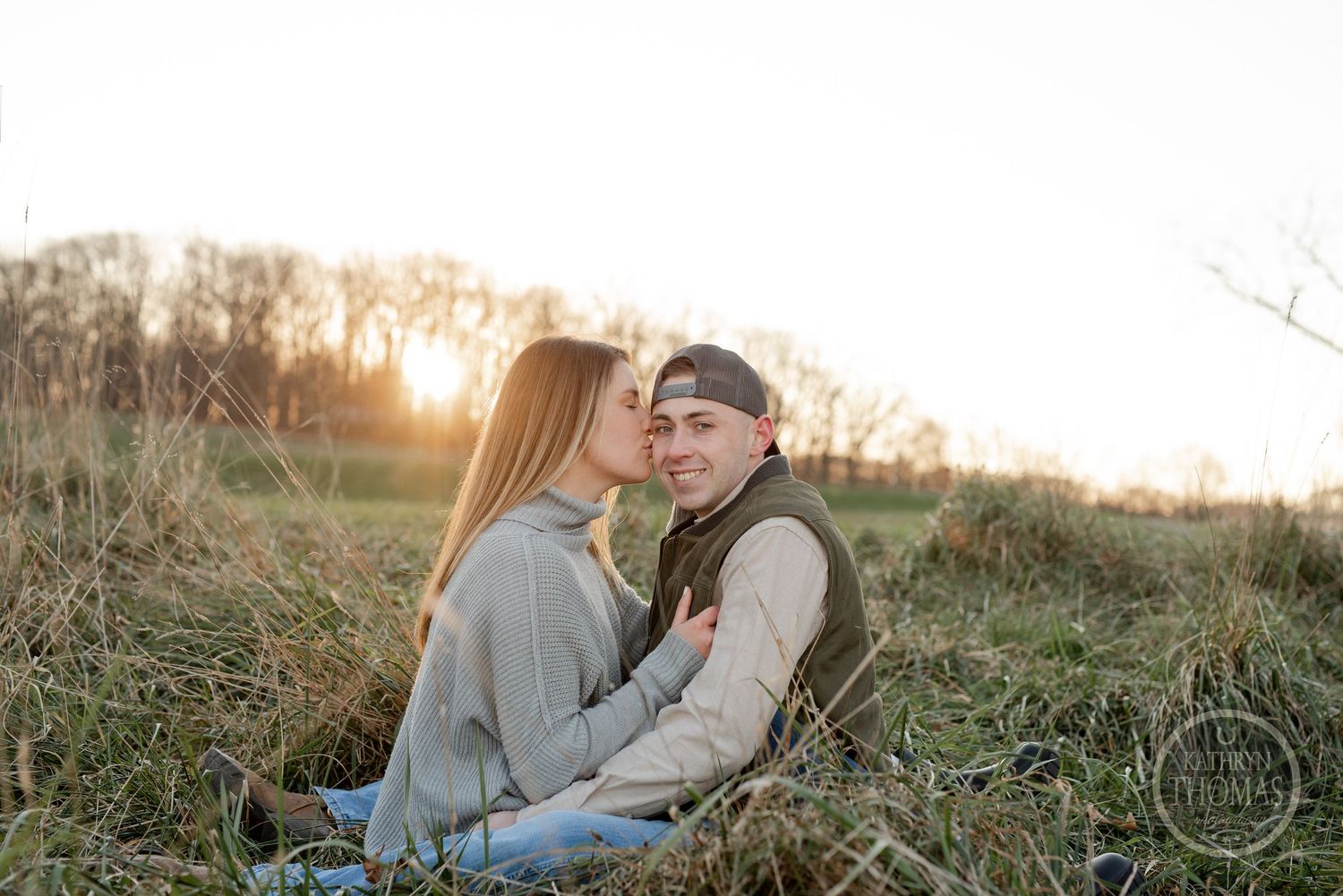 A couple embraces while sitting in a field of grass during a warm sunset.