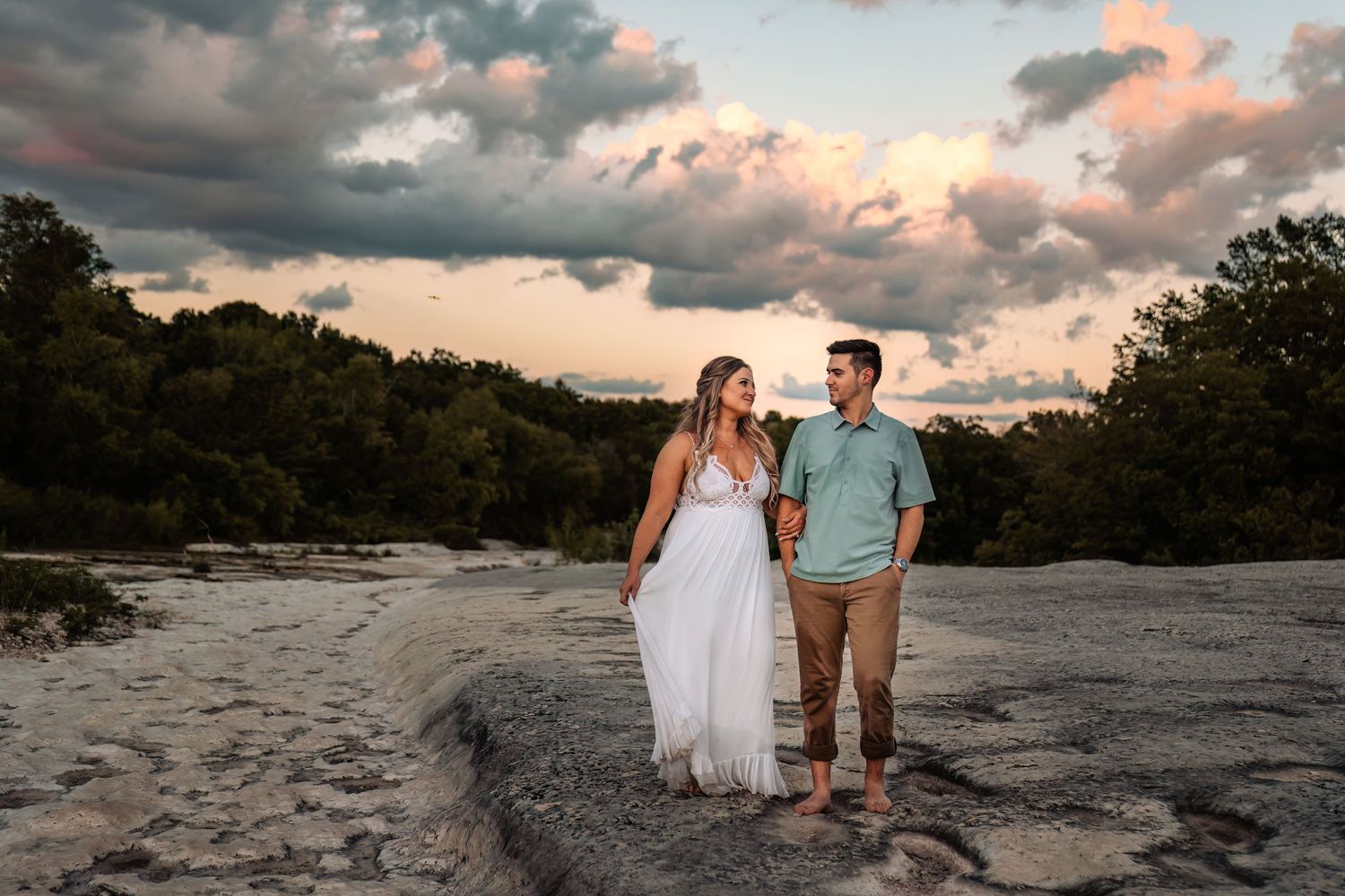 Photo of woman in long dress posing with her fiance on rocks by water by Sarah Elizabeth Photography