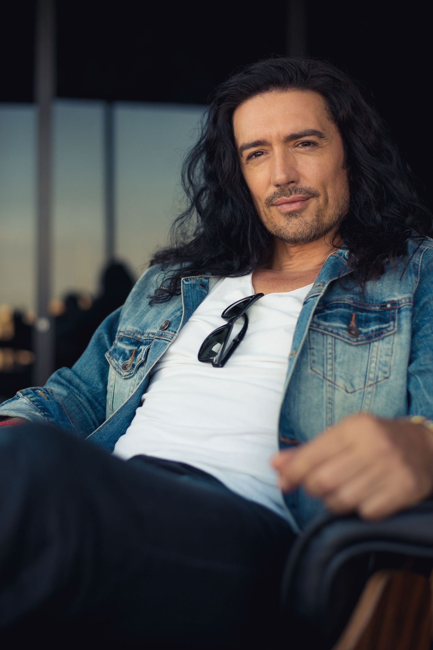 Casual portrait of person in denim jacket and white shirt with long dark hair against window.