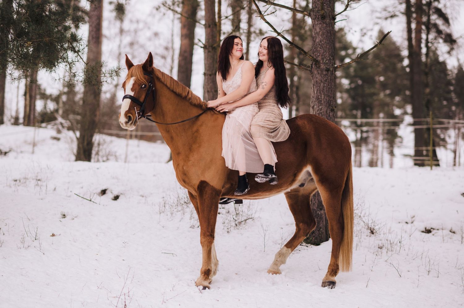 Two women sitting together on a chestnut horse in a snowy winter forest setting.
