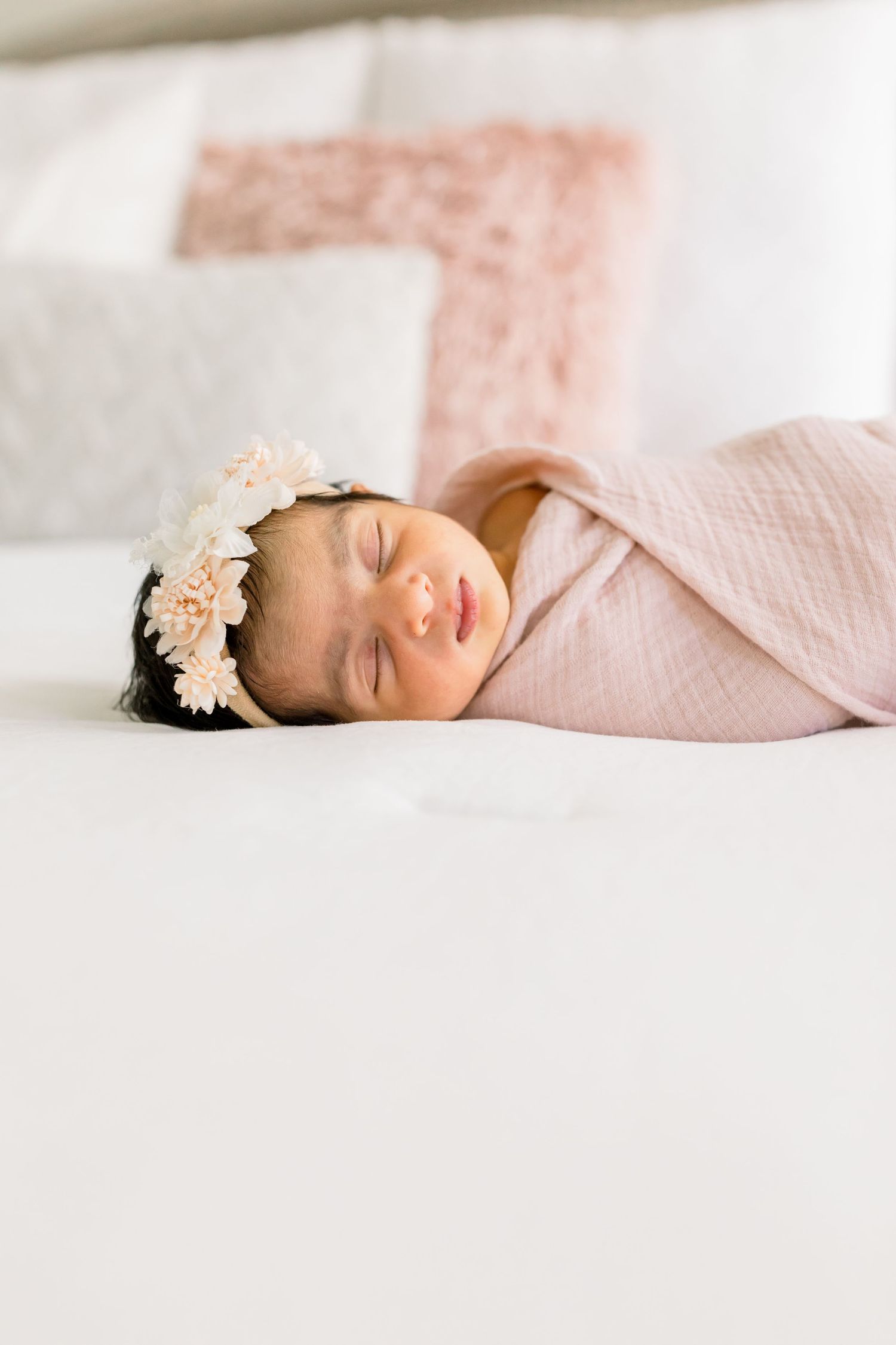 Sleeping newborn wearing floral headband and pink swaddle on white bedding for professional portrait session.