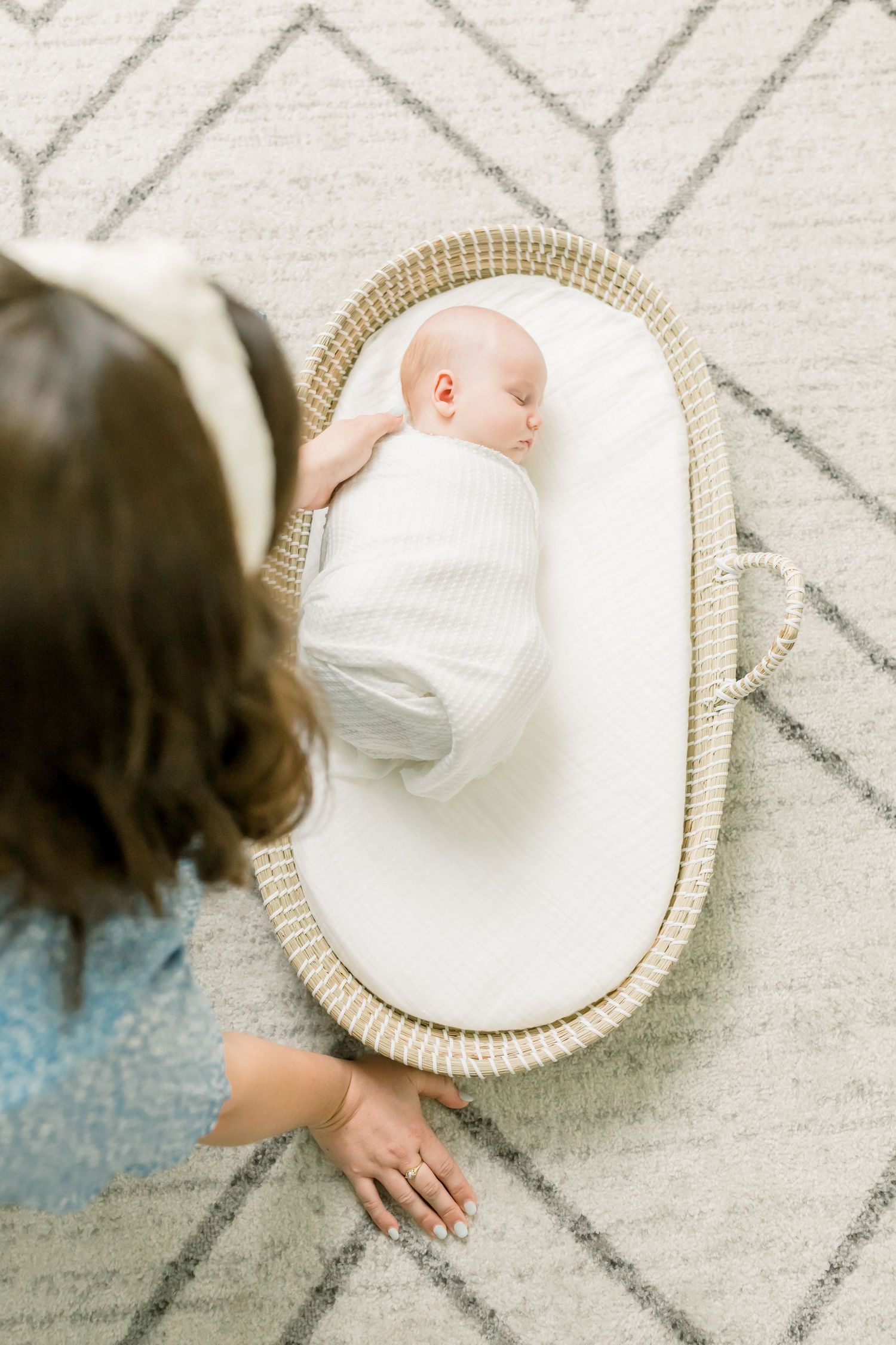 Newborn baby swaddled in white lying in a woven bassinet on geometric patterned carpet.