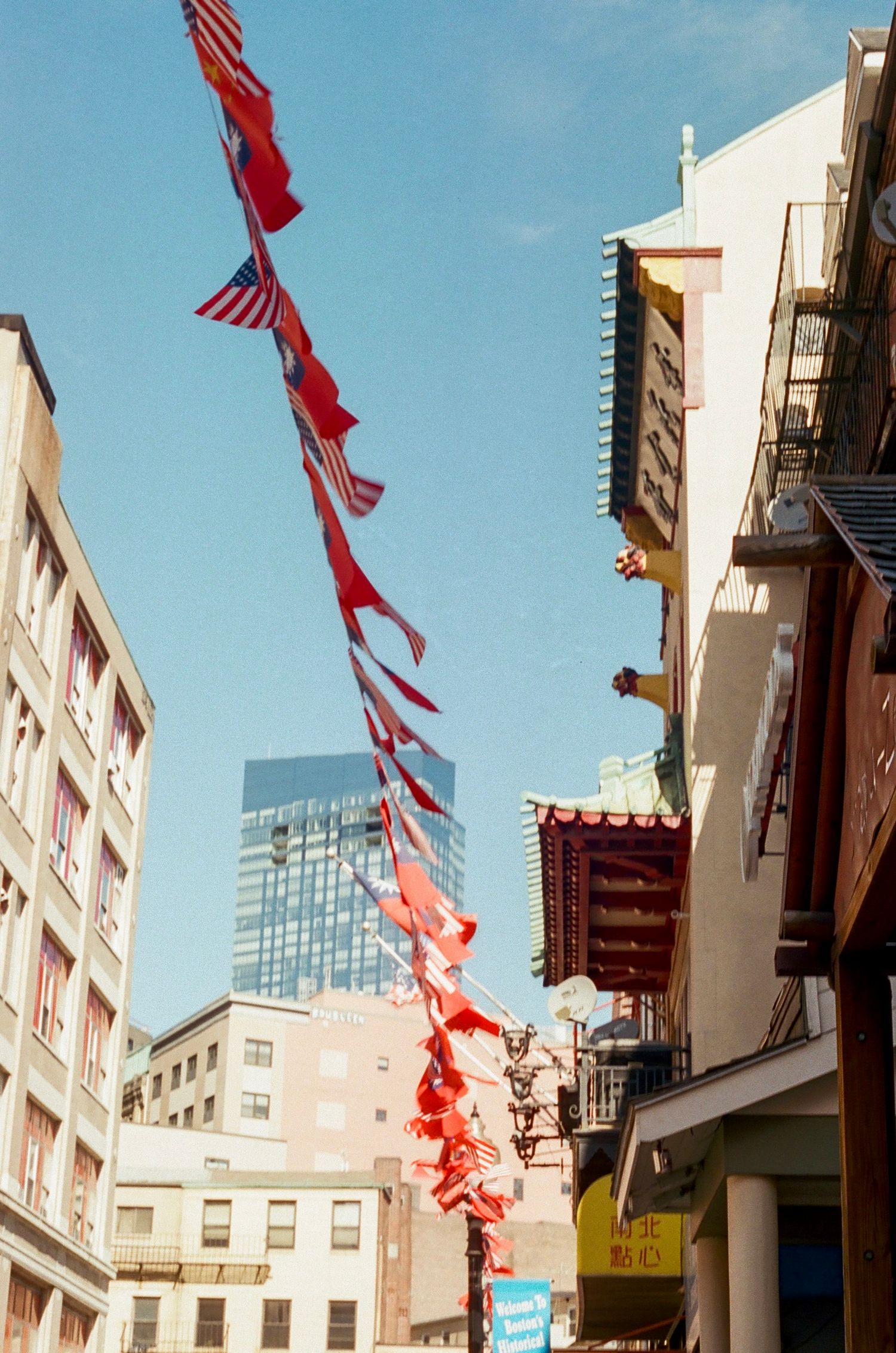 Urban street scene with red and American flags hanging from building facades against blue sky.