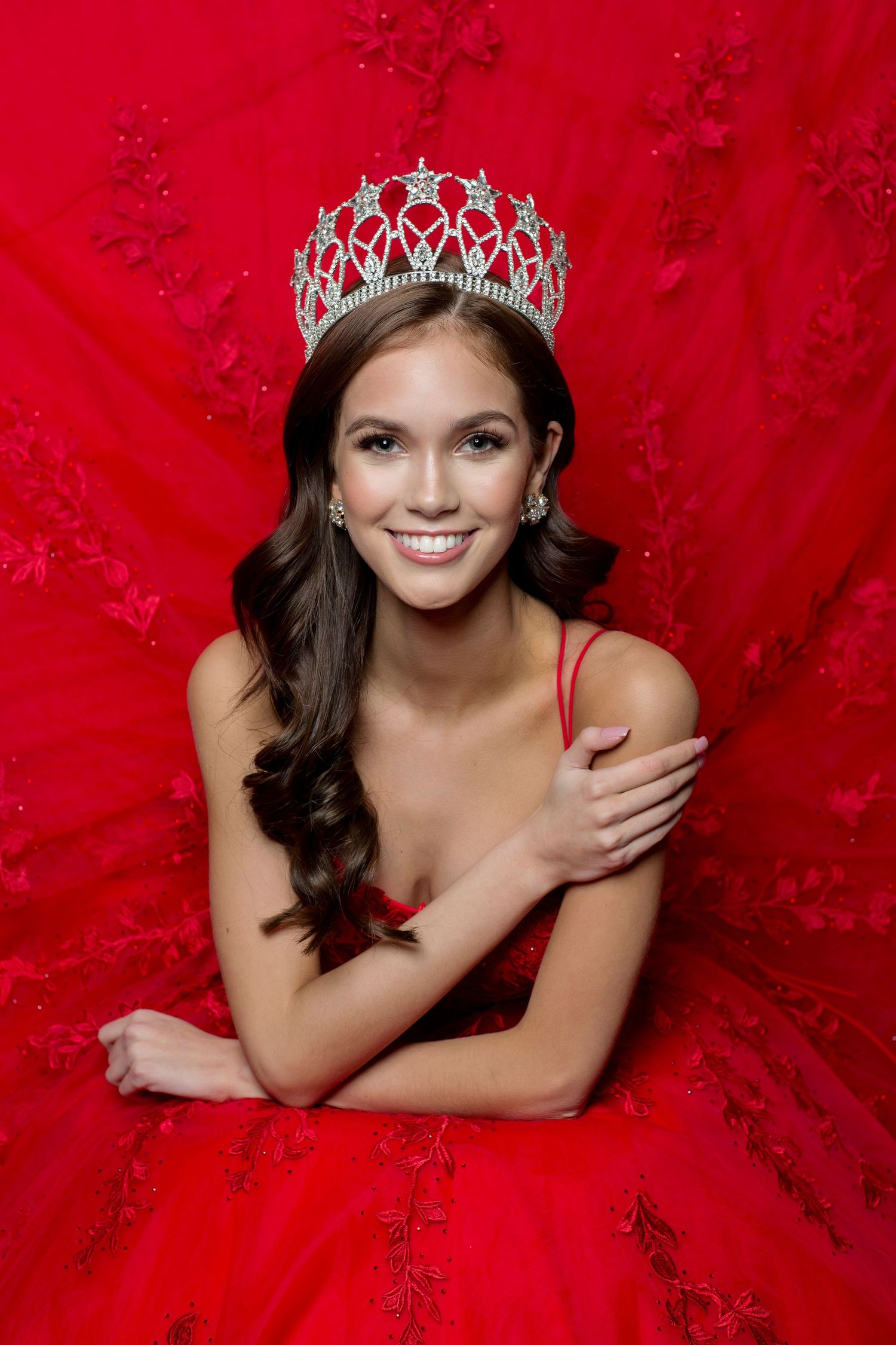 Someone wearing a sparkly silver crown and red formal gown poses for an elegant portrait against a red backdrop.
