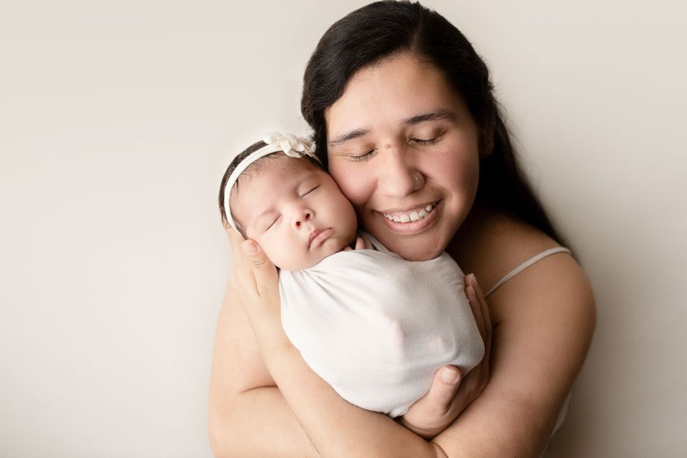 Eden Prairie newborn photographer | Hispanic-mom-holding-newborn-baby-with-bow-on-babies-head-with-cream-backdrop