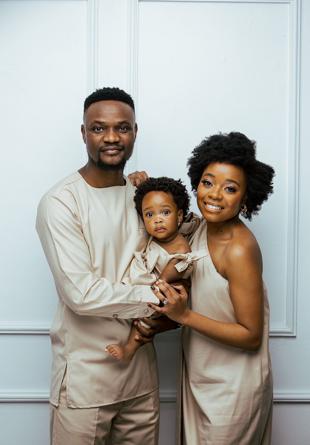 Family in coordinated neutral outfits posing together against white paneled wall.