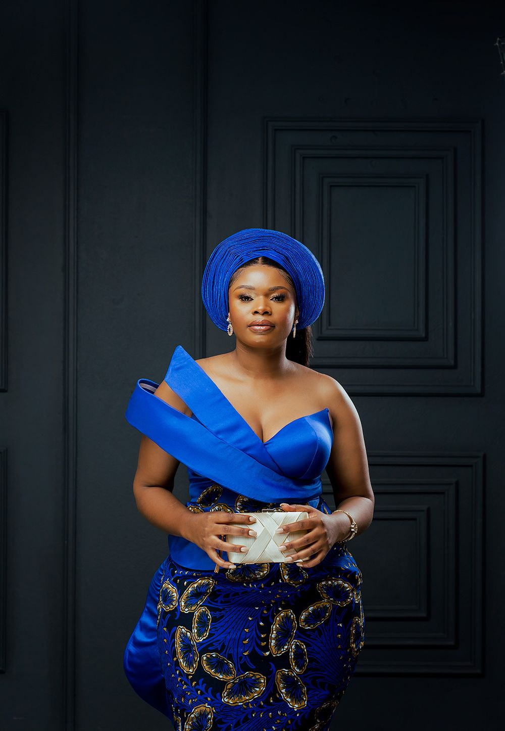 Woman in royal blue traditional dress with headwrap poses against dark paneled wall.