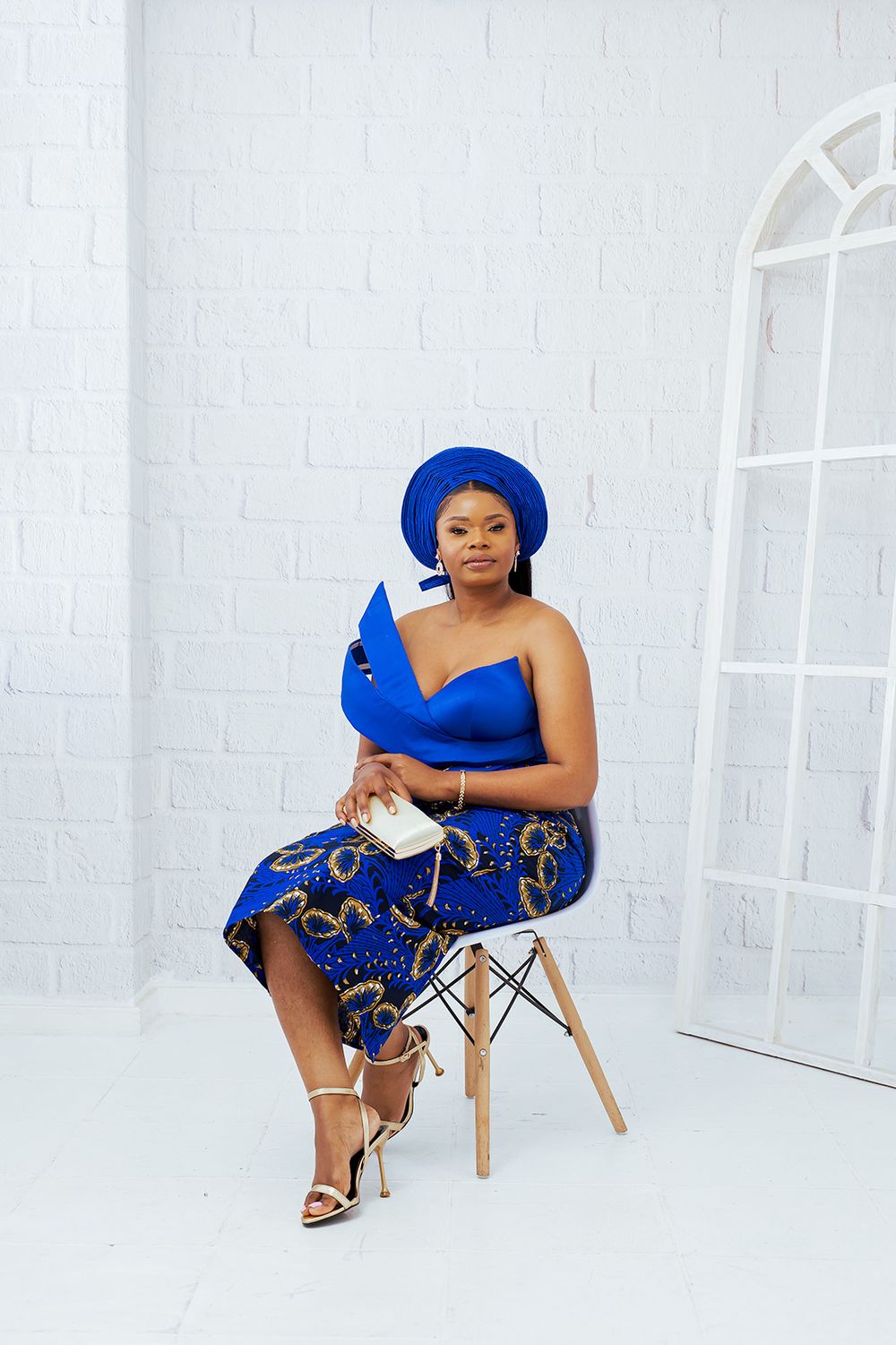 Woman in blue traditional outfit sits elegantly on chair in white studio setting.