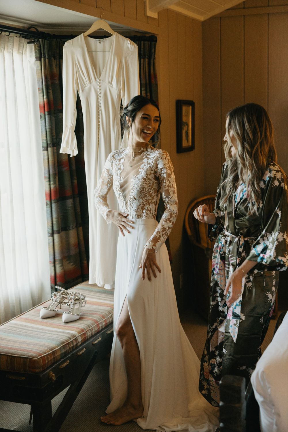 Elegant white lace wedding dress hanging in sunlit room with floral decor.