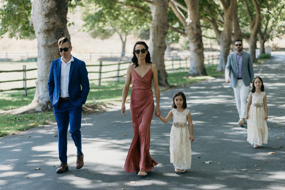 A couple and a young girl in white walk together on a tree-lined path during a sunny day.