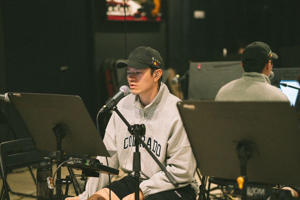 Musician in gray hoodie and cap performs with a laptop and music stand during rehearsal.