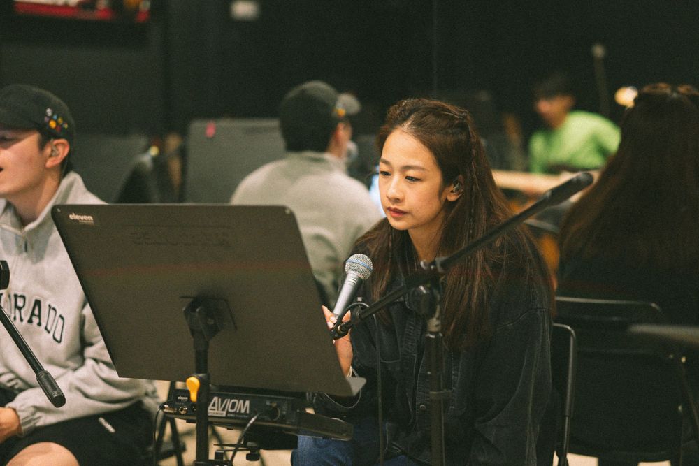 Musicians rehearse with laptops and microphones in a dimly lit studio space.