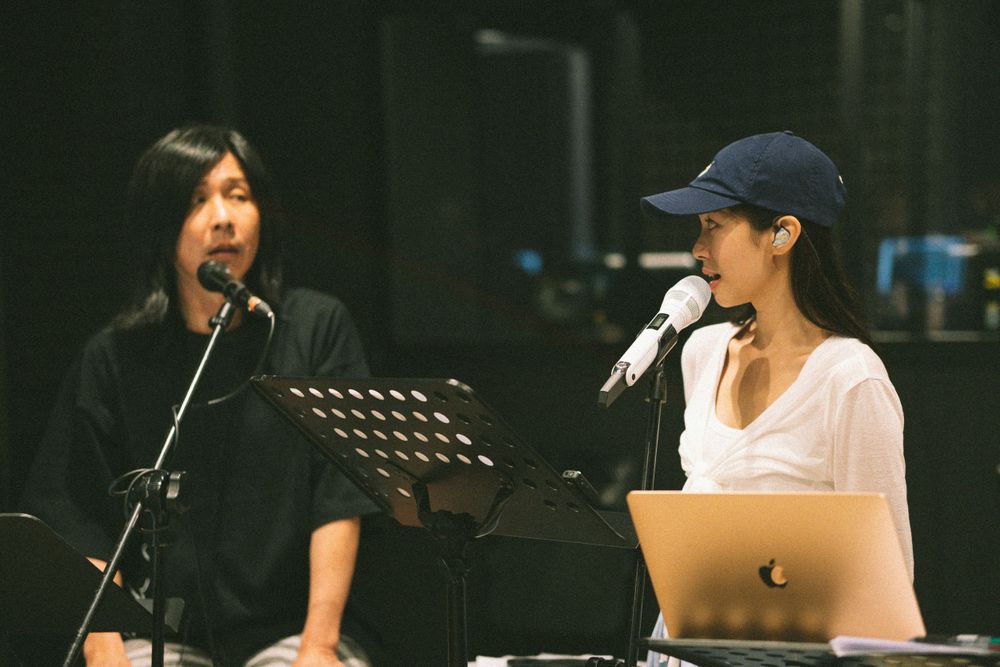 Musicians perform together with microphones and music stands on dark stage.
