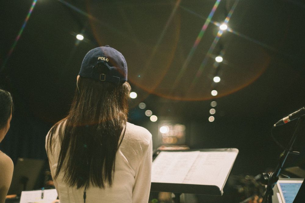 Back view of performer with long hair wearing baseball cap during studio rehearsal.